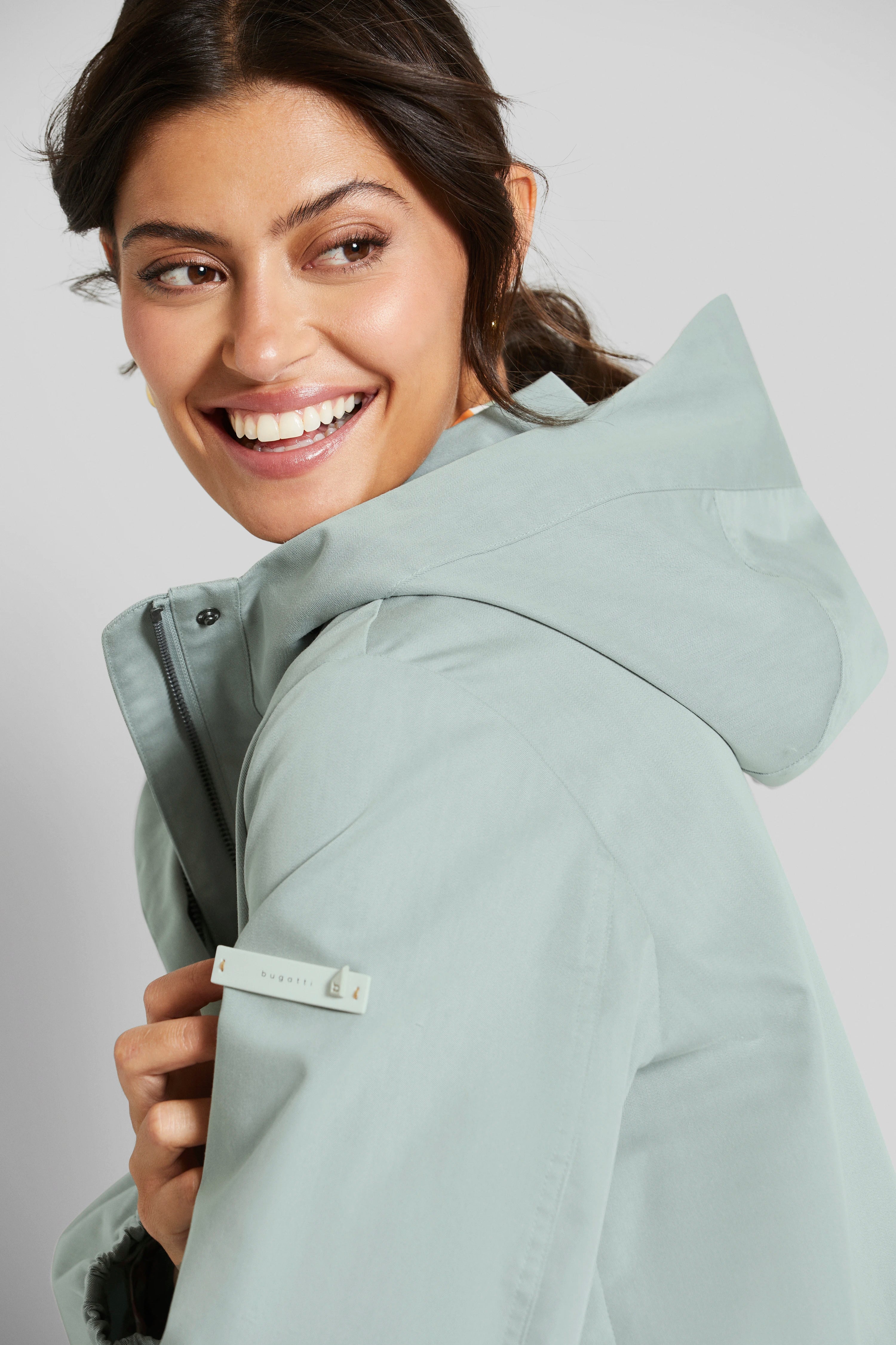 Modelbild in grauer bugatti-Jacke mit Kapuze, lächelndes Posing.