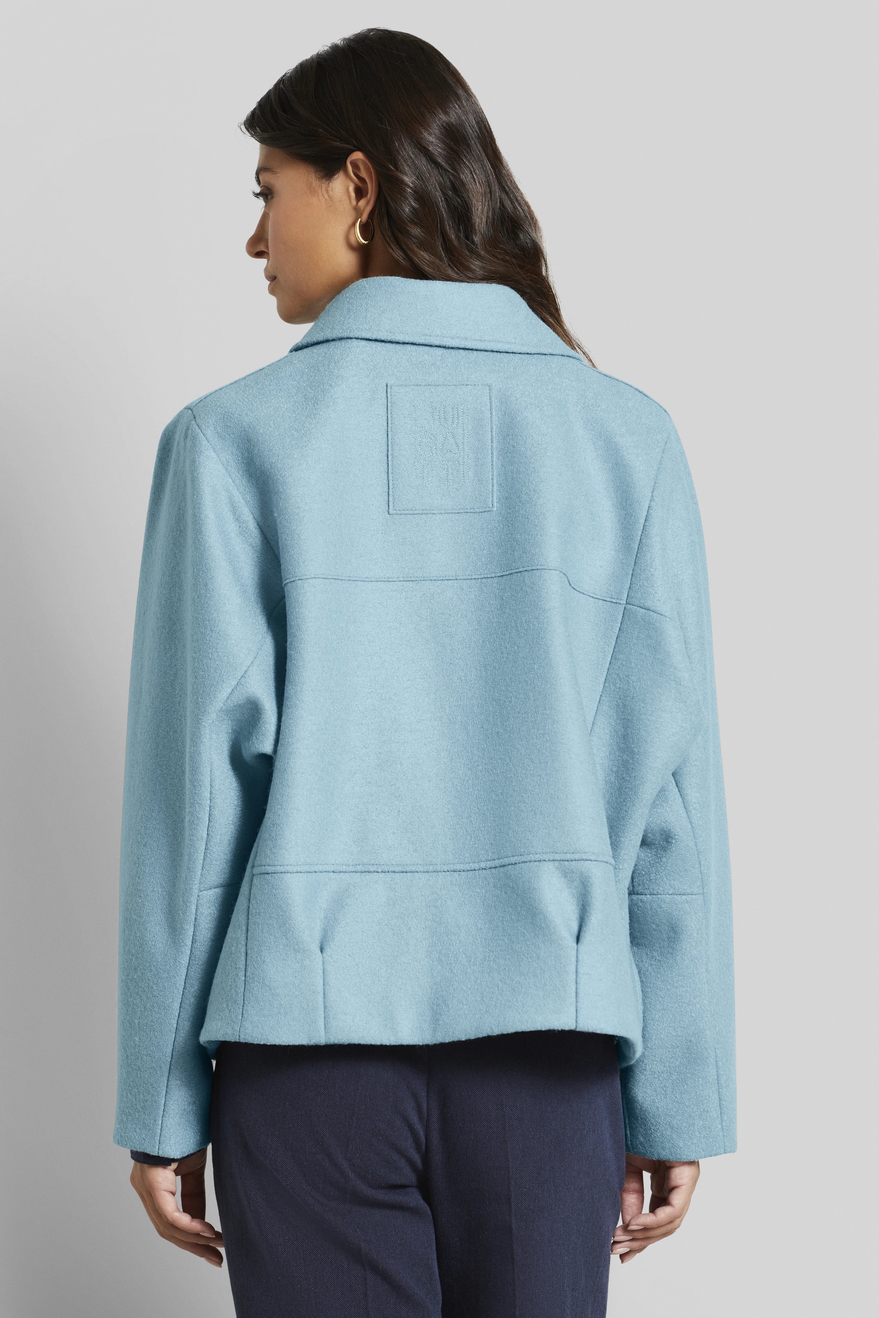 Model draagt lichtblauwe bugatti mantel, achterkant, ontspannen pose, gecombineerd met donkerblauwe broek.
