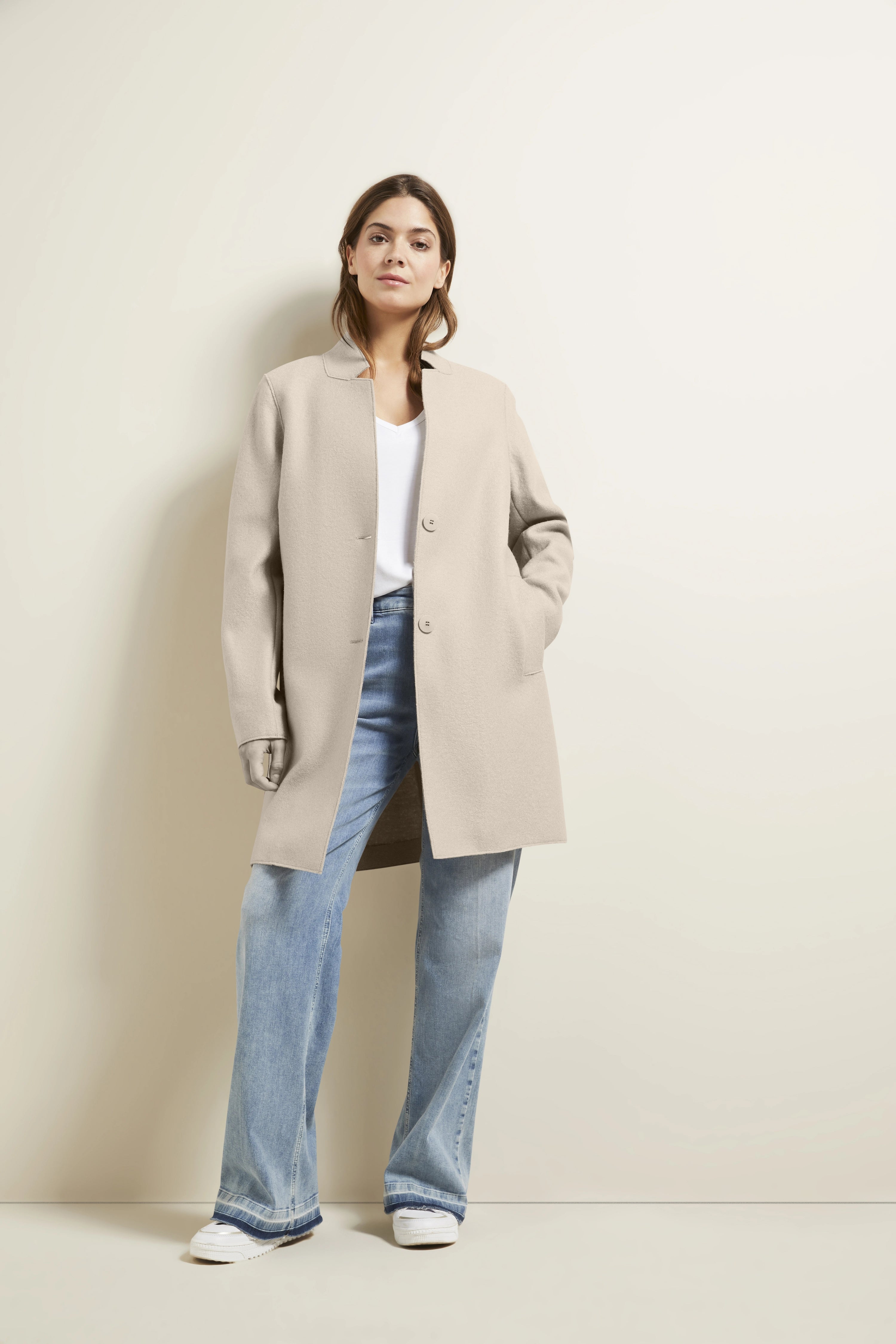 Model in beigem bugatti-Mantel, weißem Shirt, blauen Jeans, lässig posierend.