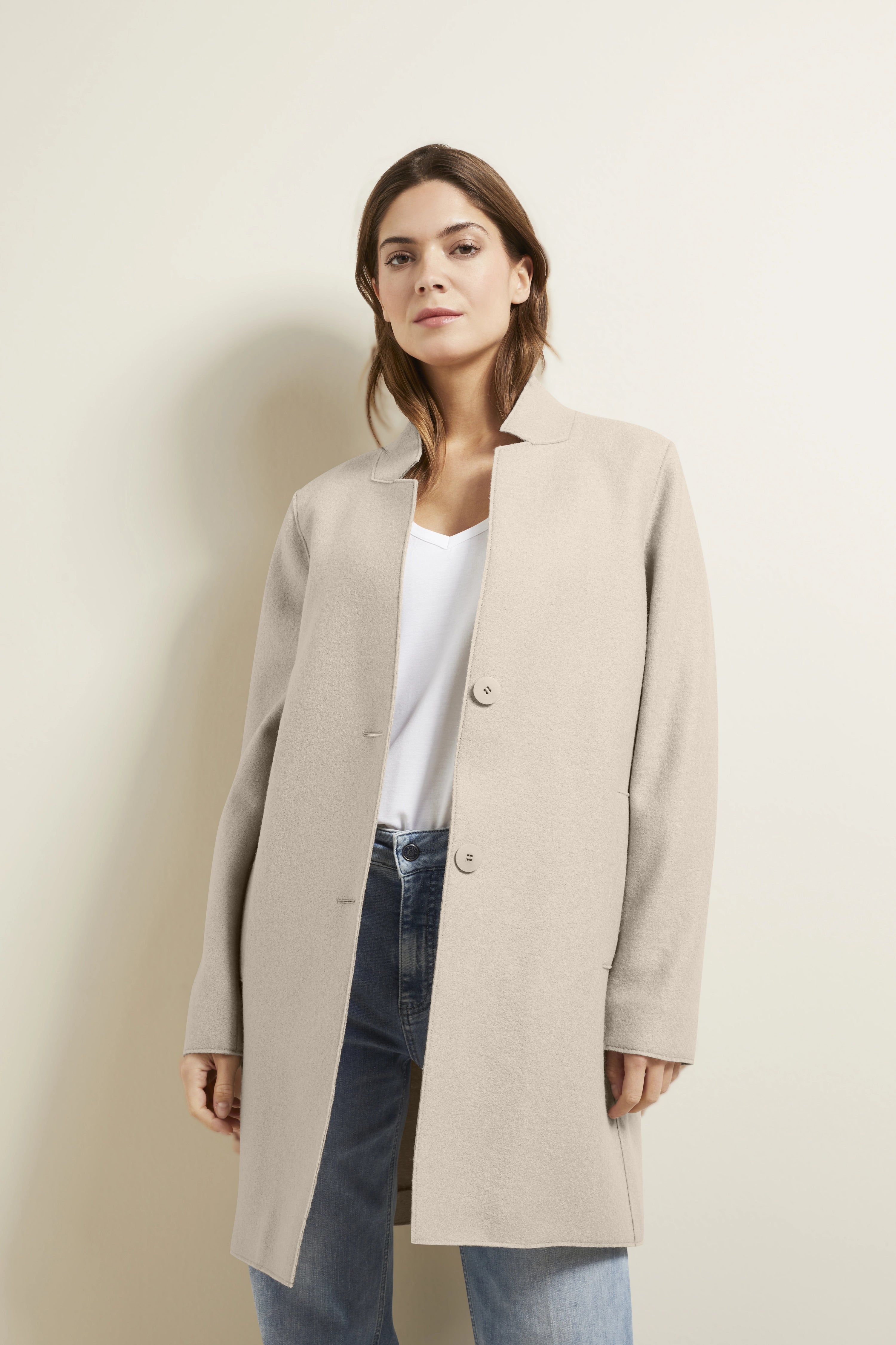 Beiger bugatti Mantel auf Model, weißes Shirt, blaue Jeans, entspannte Pose.