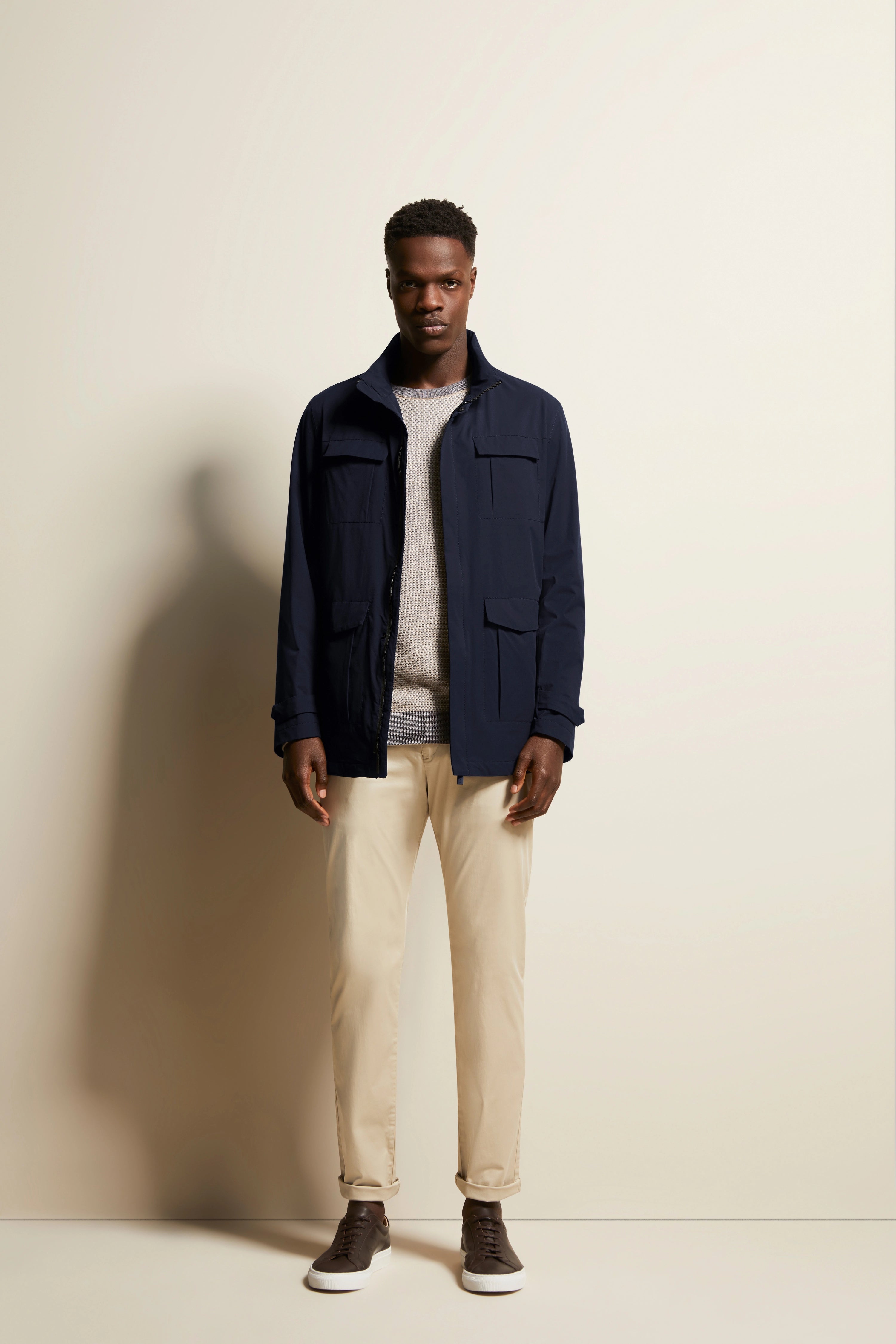 Modelbild: Navy Jacke, grauer Pullover, beige Chinos, braune Sneakers, entspanntes Posing.