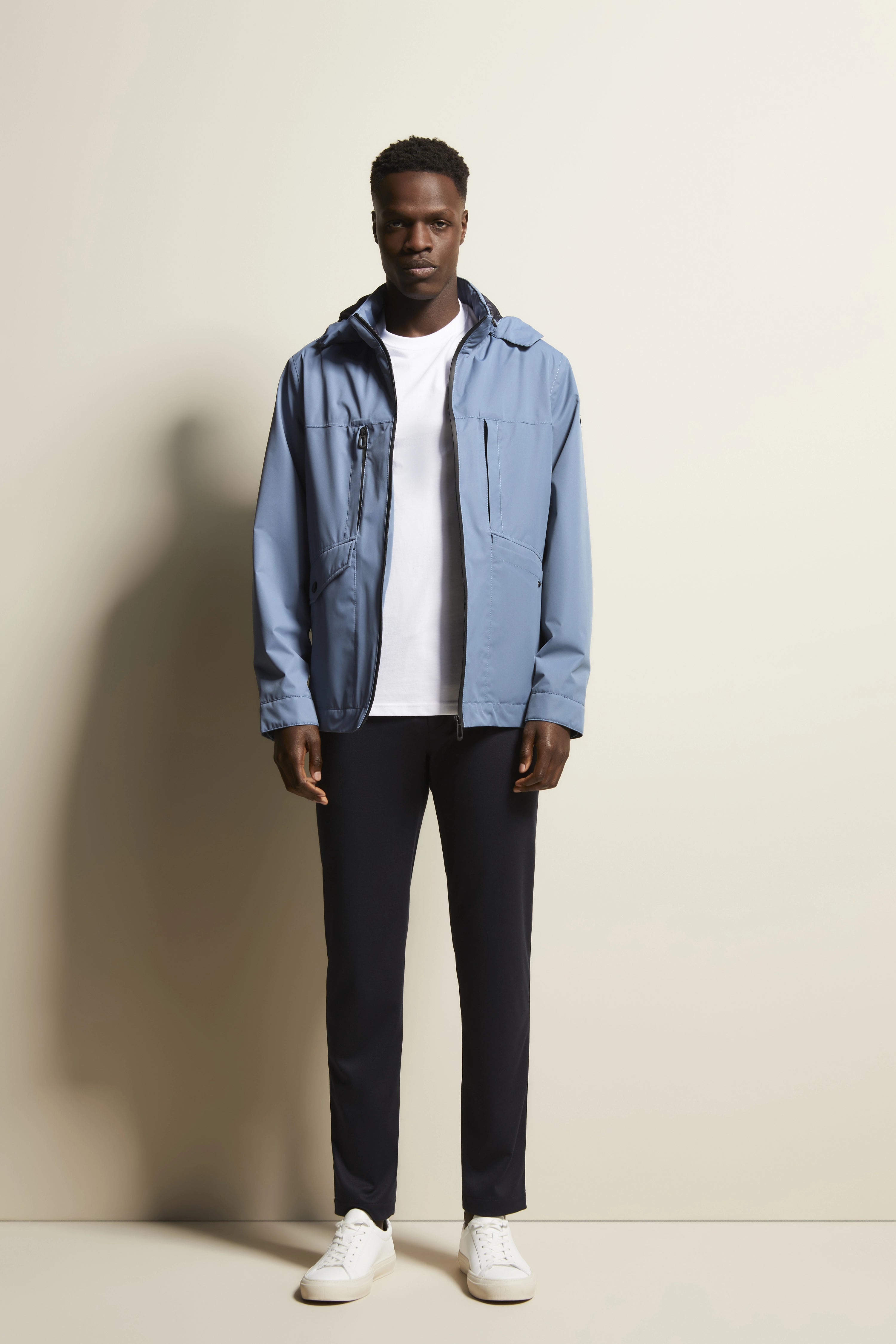 Model in blauer bugatti Jacke, weißem Shirt, dunkler Hose und weißen Sneakers, frontal stehend.