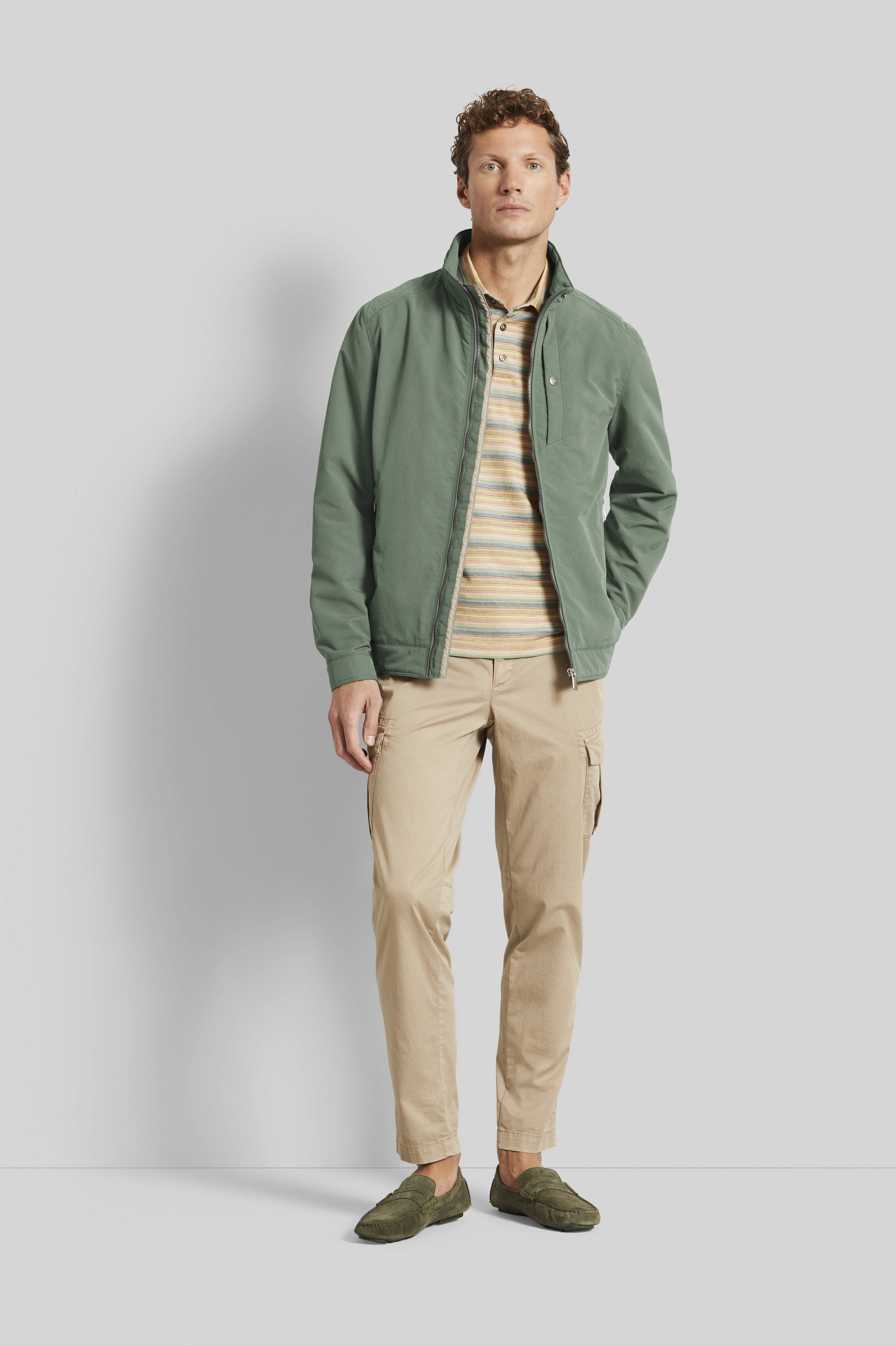 Model in green bugatti blouson, striped polo, beige cargo pants, green shoes, standing casually.