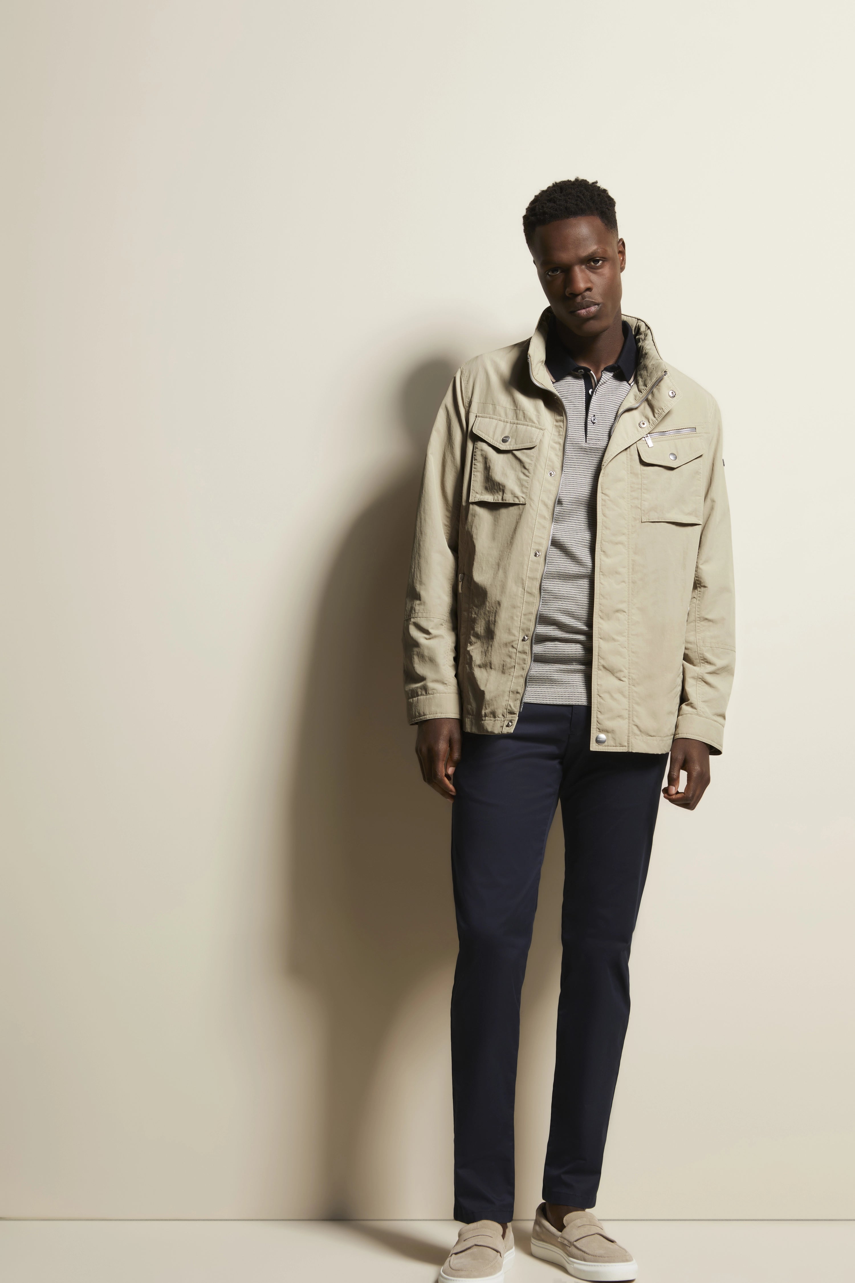 Model in beiger Jacke, grauem Polo, dunkelblauer Hose und beigen Schuhen, legeres Posing.