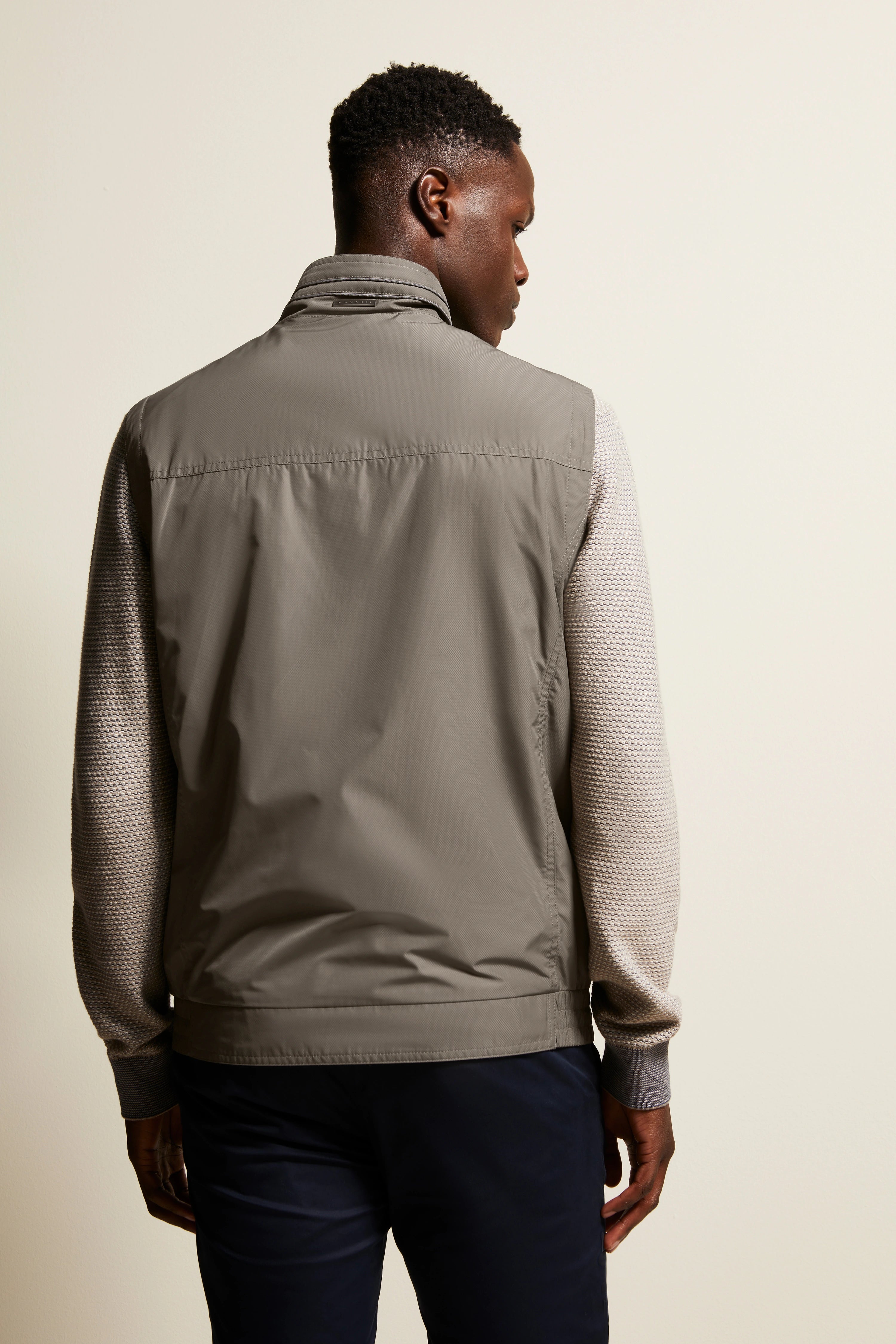 Back view: Model in gray vest, beige knit sleeves, dark pants, relaxed pose.