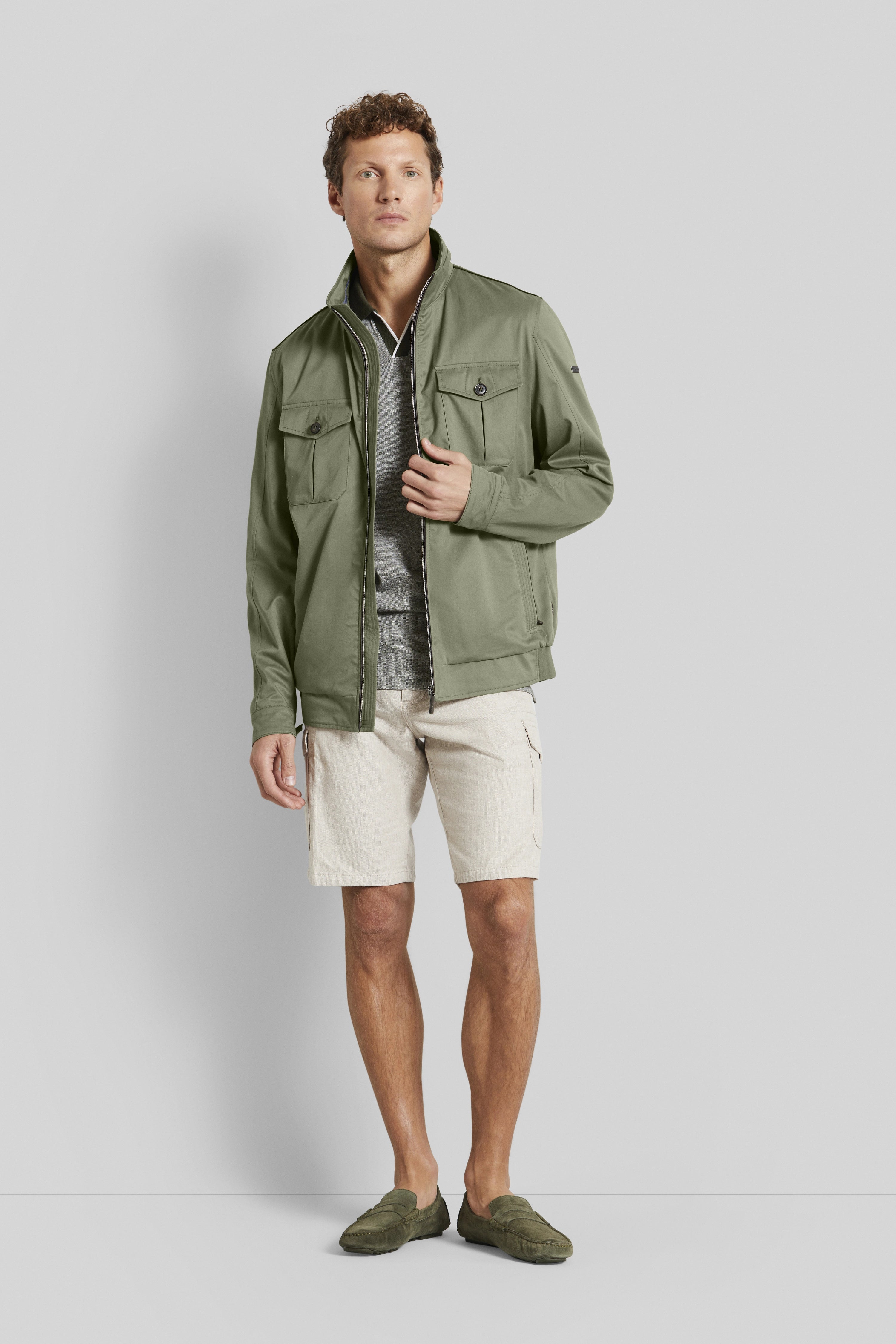 Model in olivgrüner Jacke, grauem Shirt, beiger Shorts und grünen Loafers von bugatti, lässiger Stand.