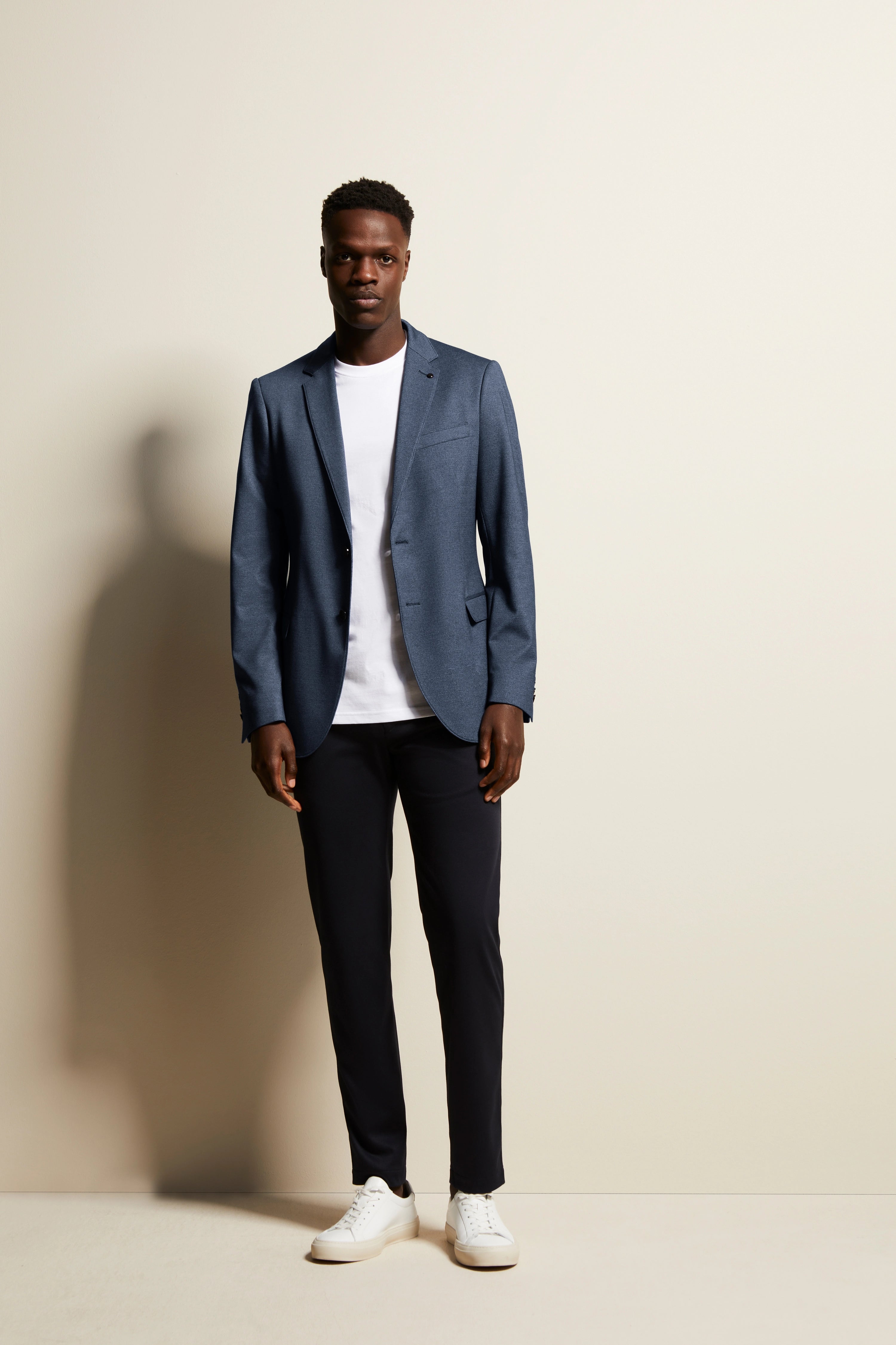 Model in bugatti donkerblauwe blazer, wit T-shirt, zwarte broek, witte sneakers. Ontspannen, rechtopstaand poseren.