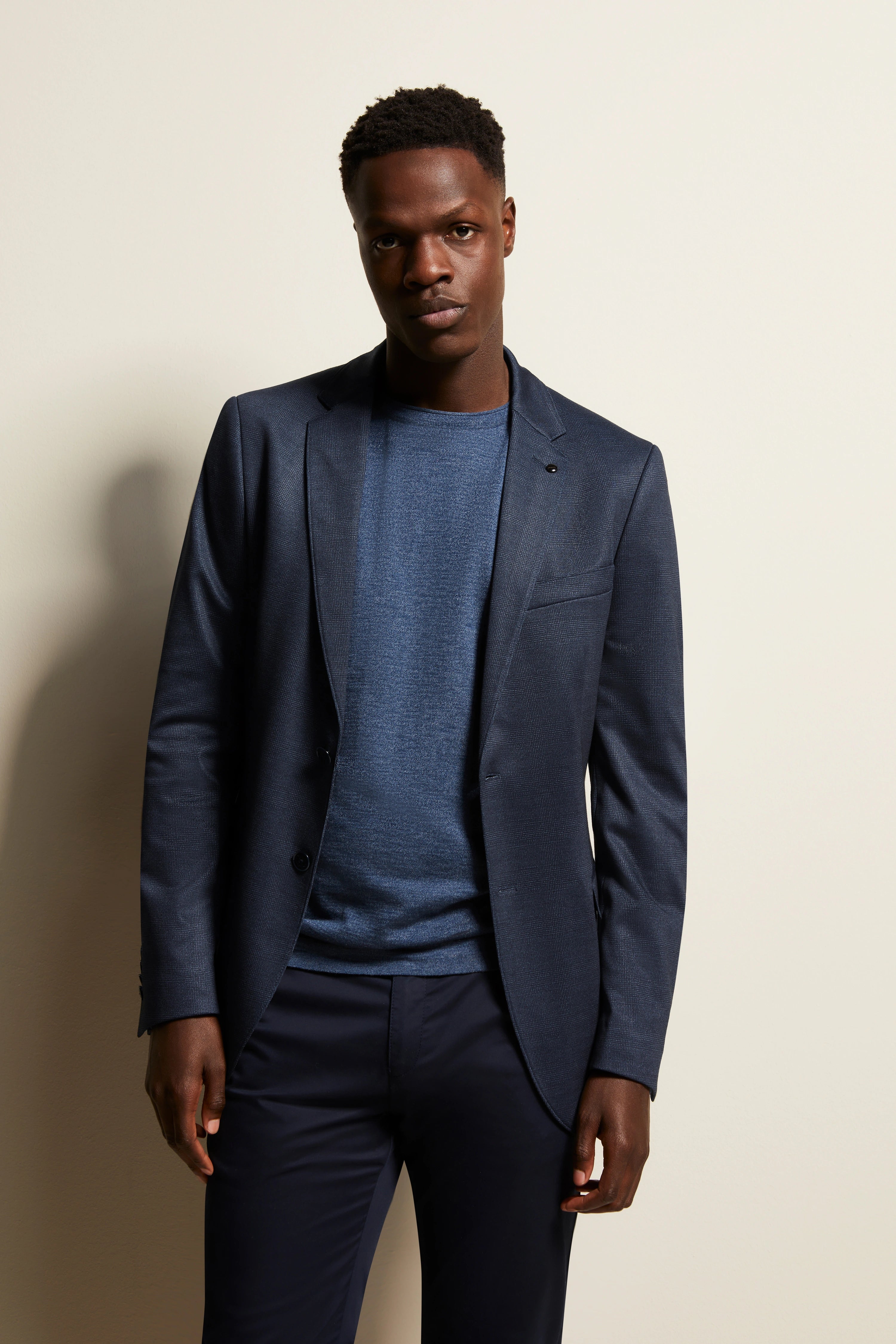 Elegantes Modelbild: Mann in navy bugatti Anzug und blauem Pullover, entspannte Pose vor hellem Hintergrund.