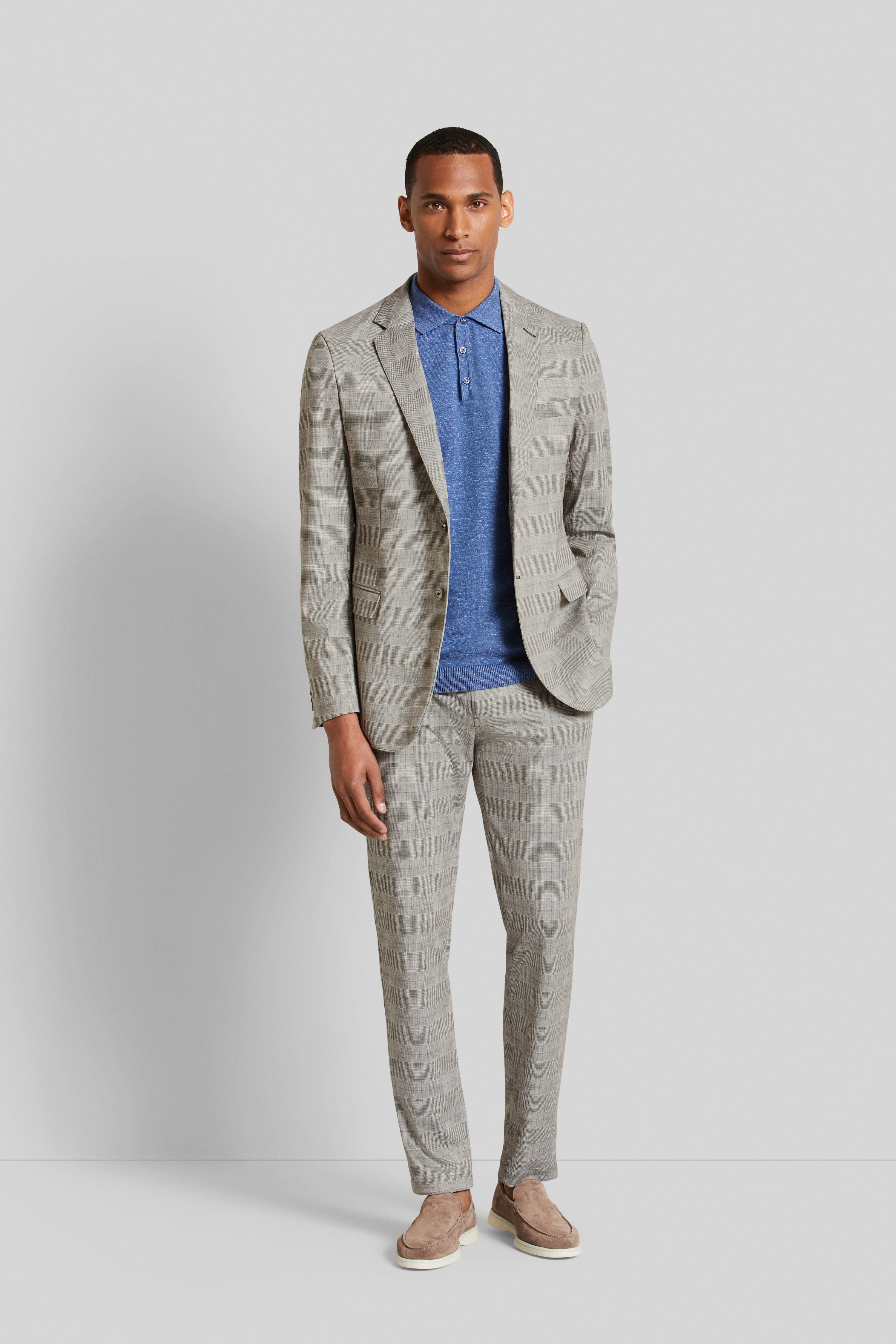 Model in grijs geruit colbert, blauwe polo, beige loafers. Staat ontspannen, handen in zakken.