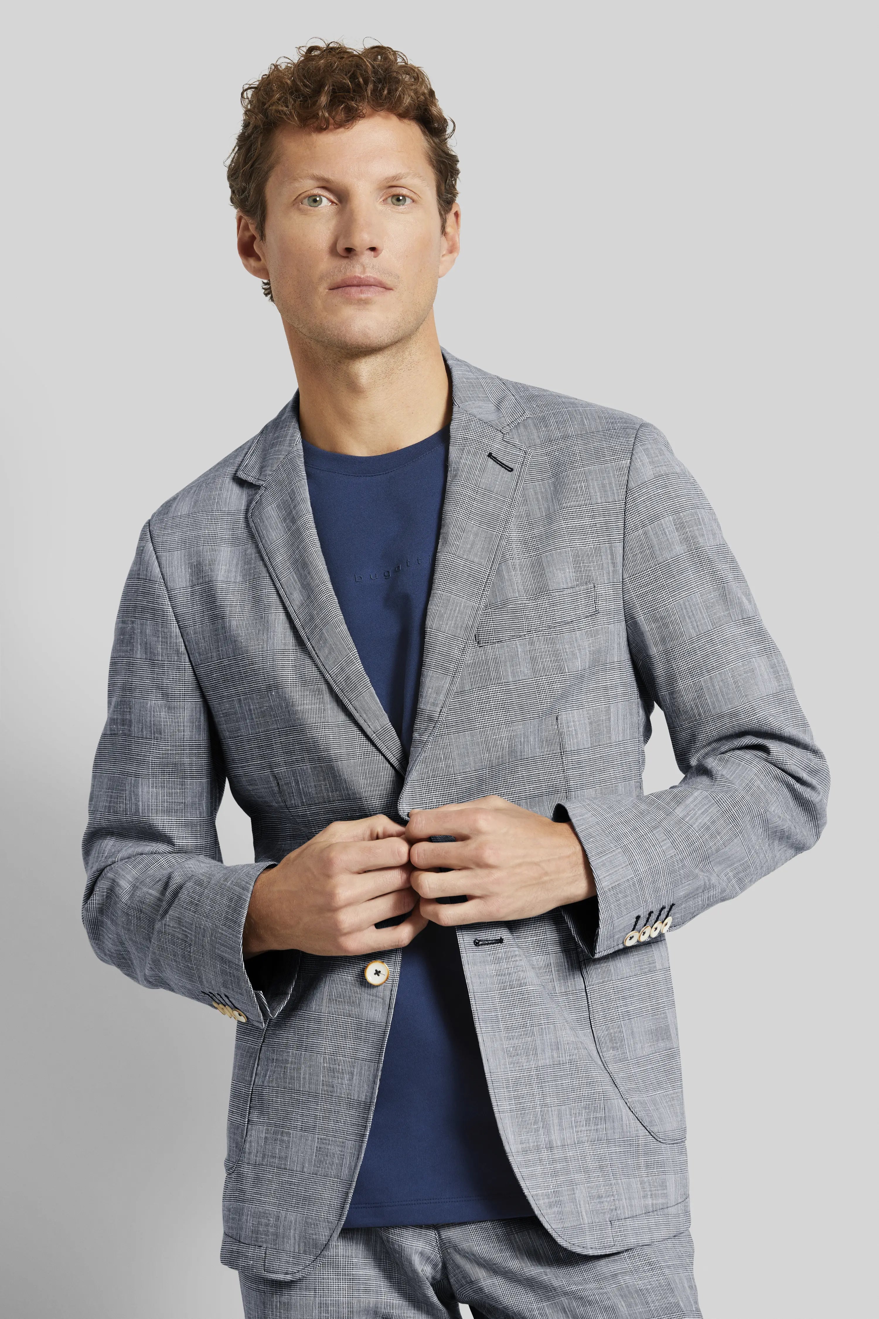 Model in gray checkered bugatti suit with blue shirt, elegant posing, hands on button.