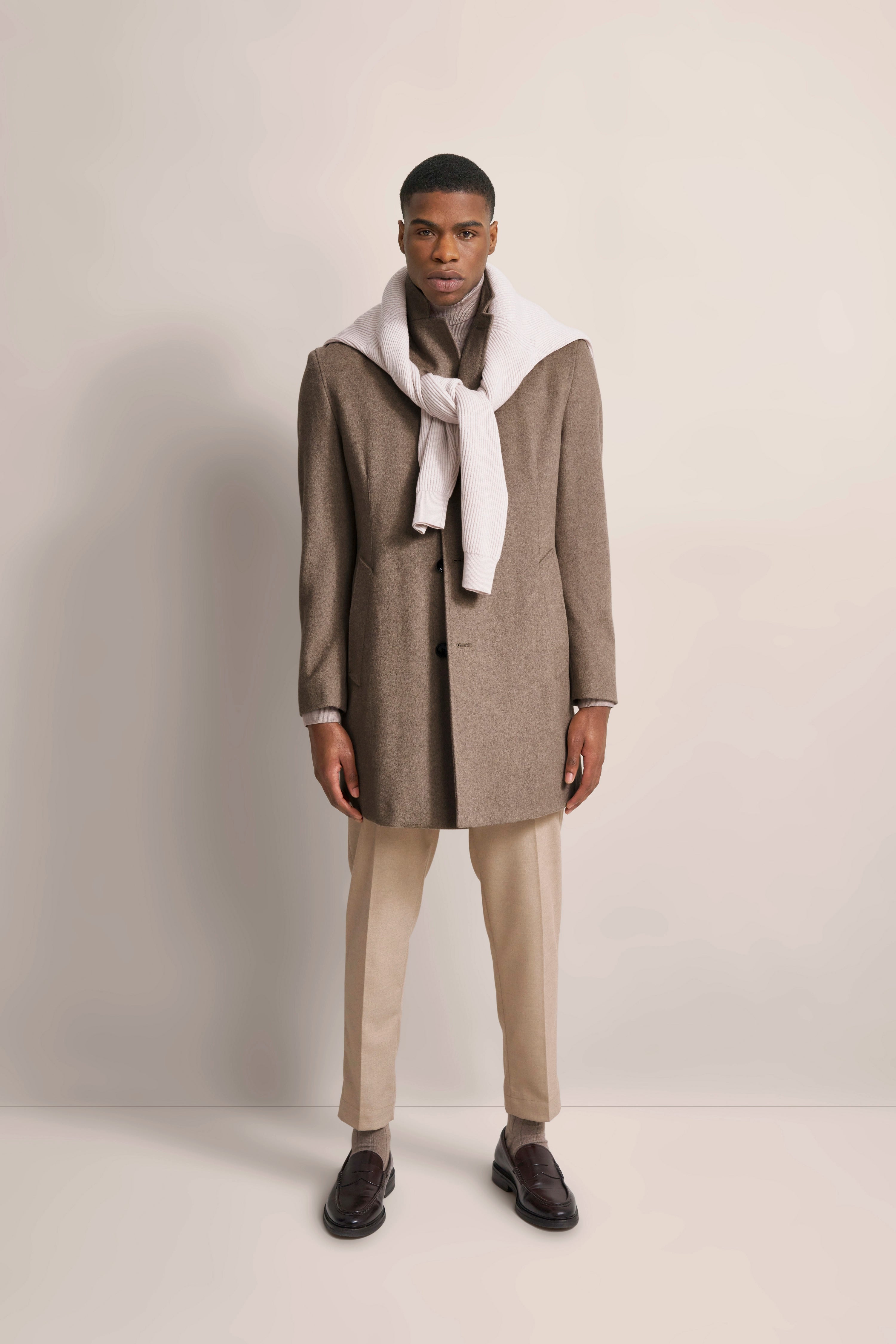Elegant model in brown coat, beige pants, sweater draped over shoulders, dark shoes by bugatti.