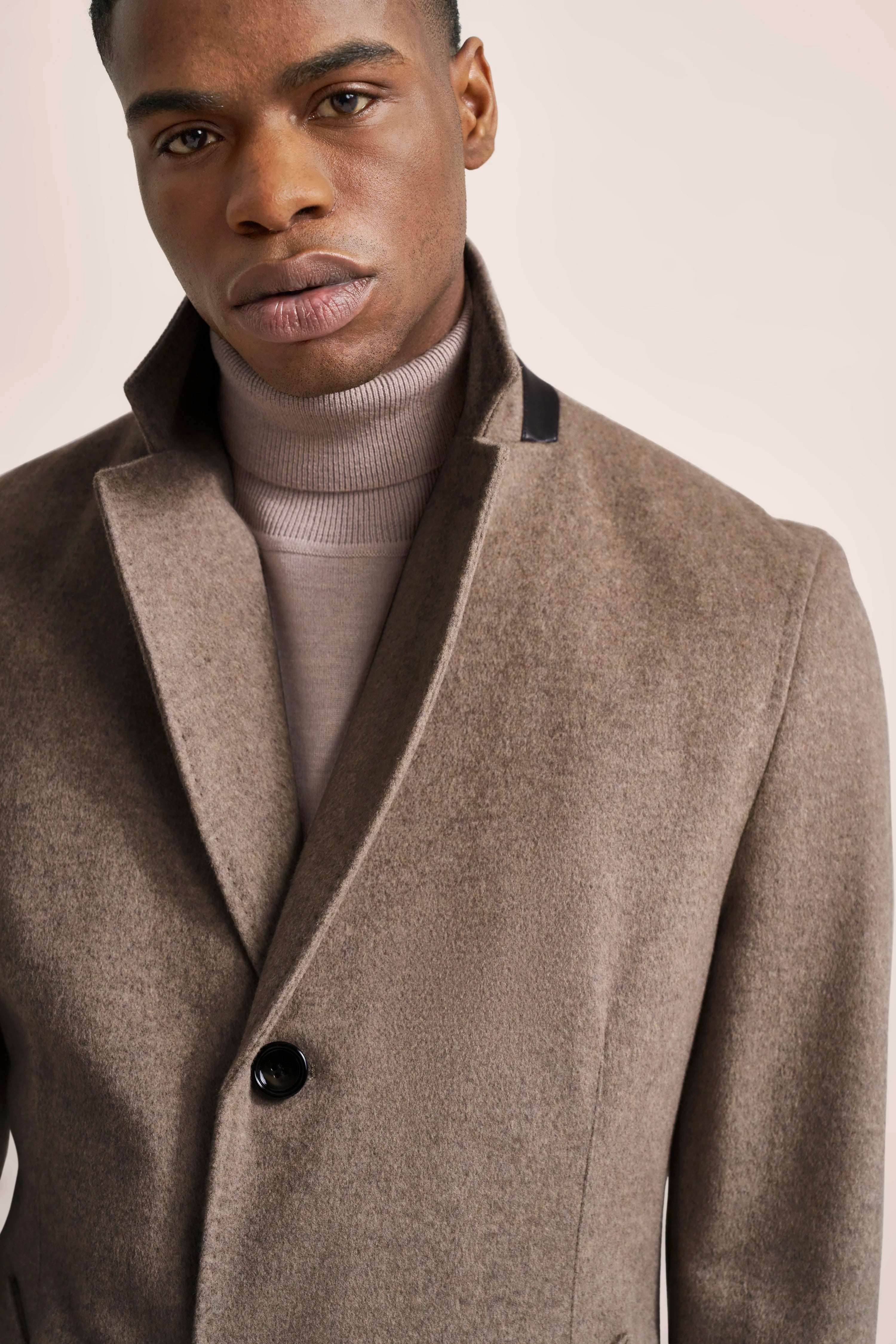 Brown coat with stand-up collar over beige turtleneck sweater, model's upper body front view.