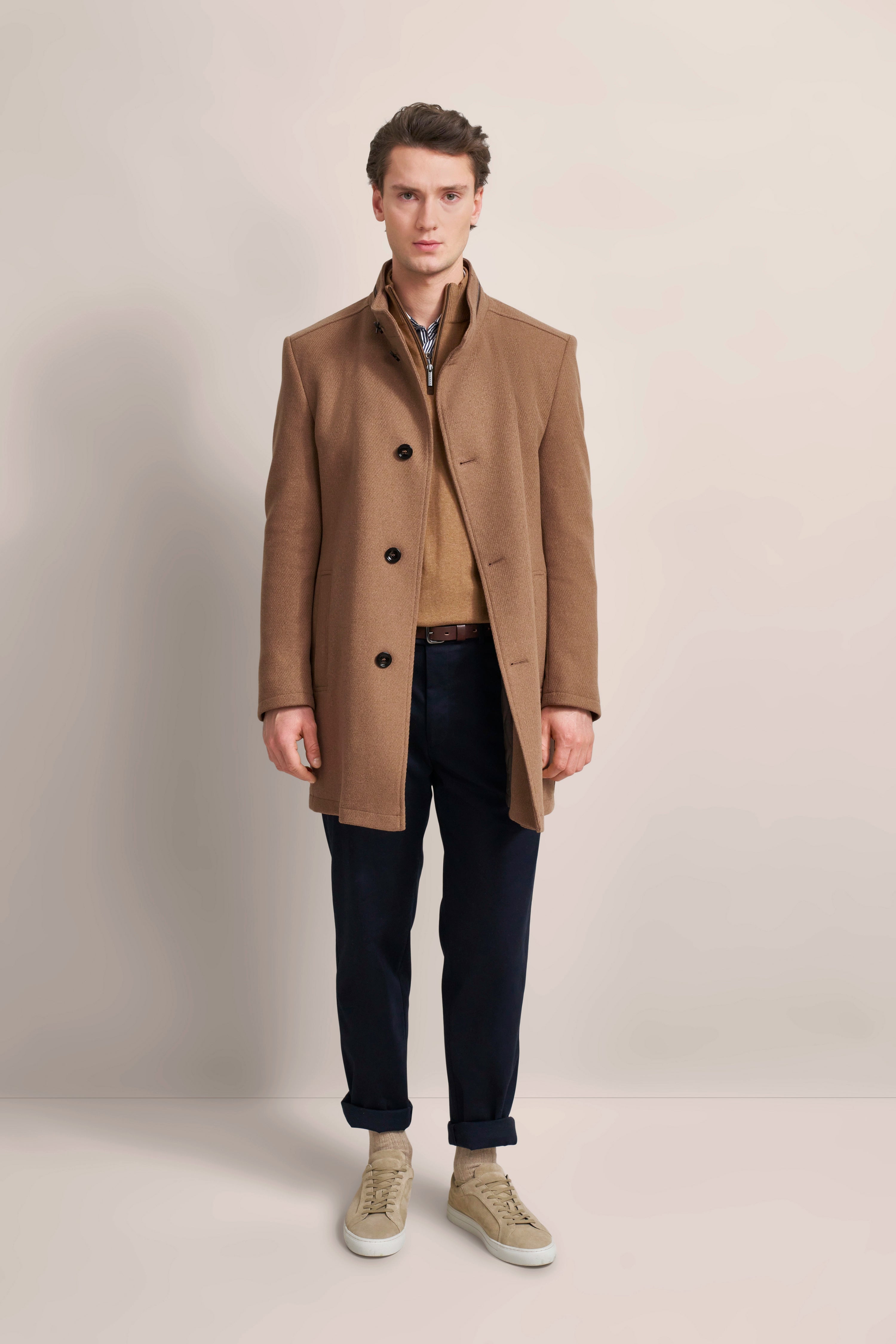 Model in camel coat, beige sweater, navy trousers, light beige sneakers. Standing pose. bugatti elegance and style.