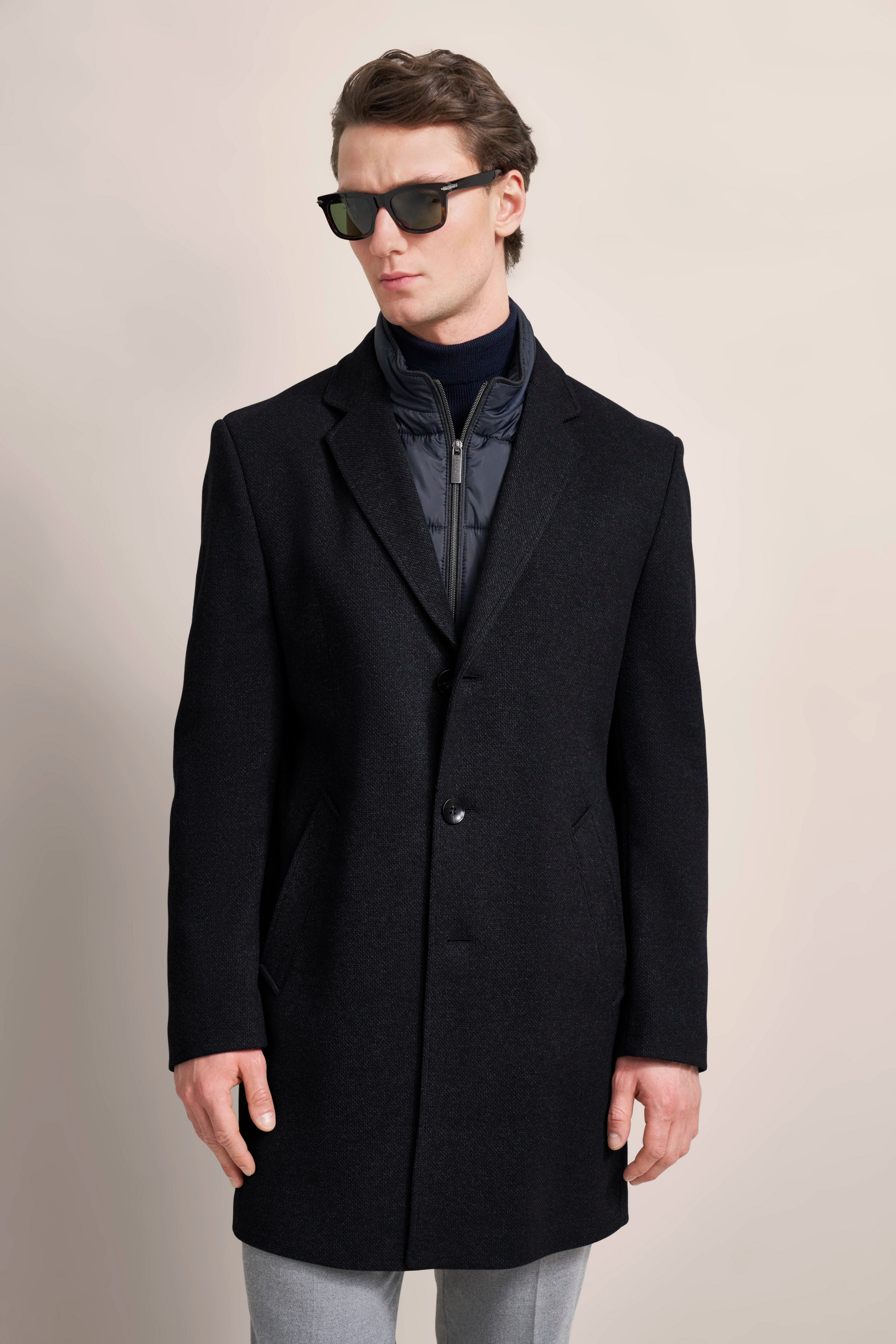 Model im schwarzen bugatti Mantel, graue Hose, Sonnenbrille, entspannte Pose.