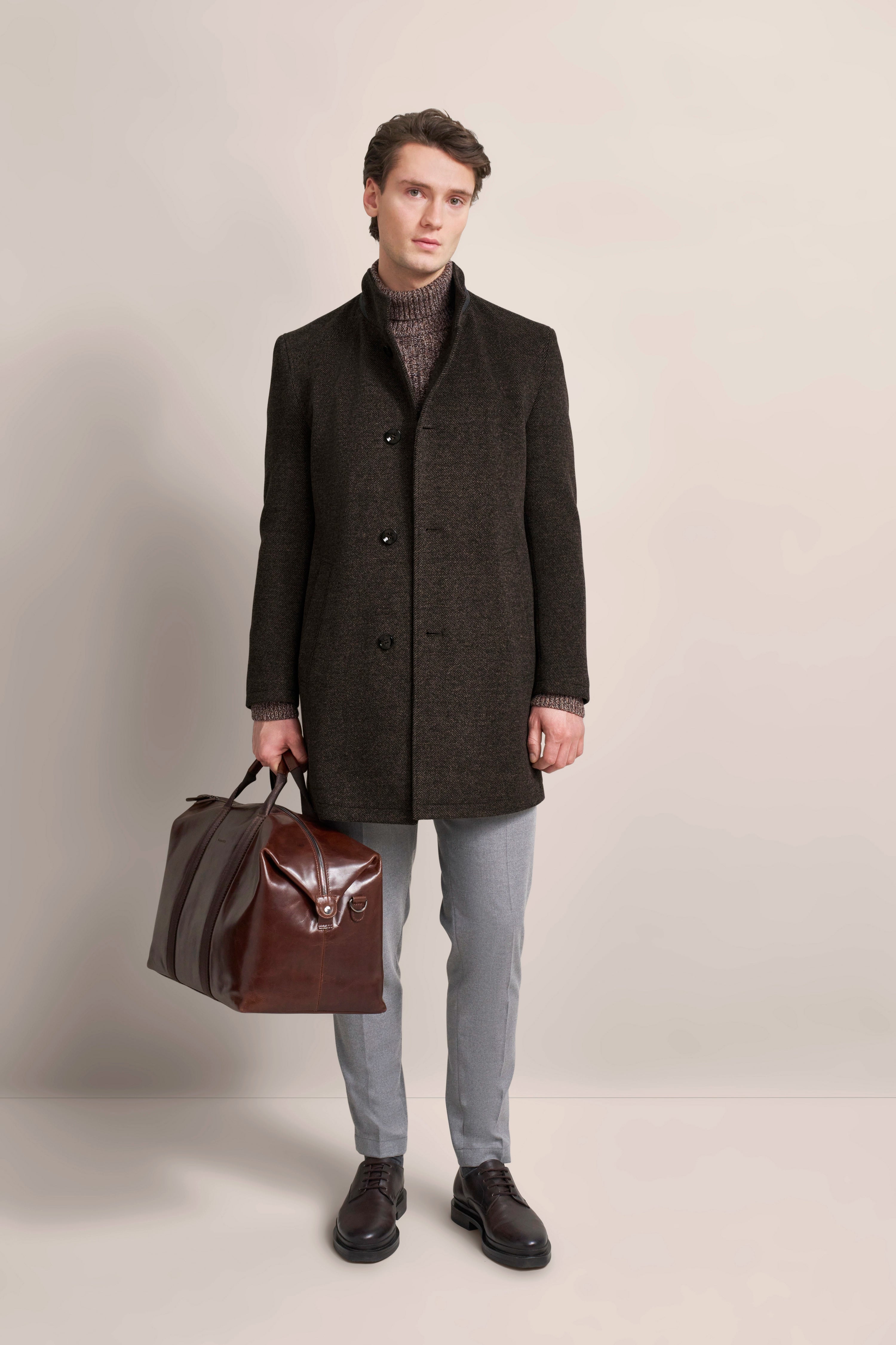 Elegantes bugatti Modelbild: Dunkler Mantel, graue Hose, braune Tasche, legeres Posing.