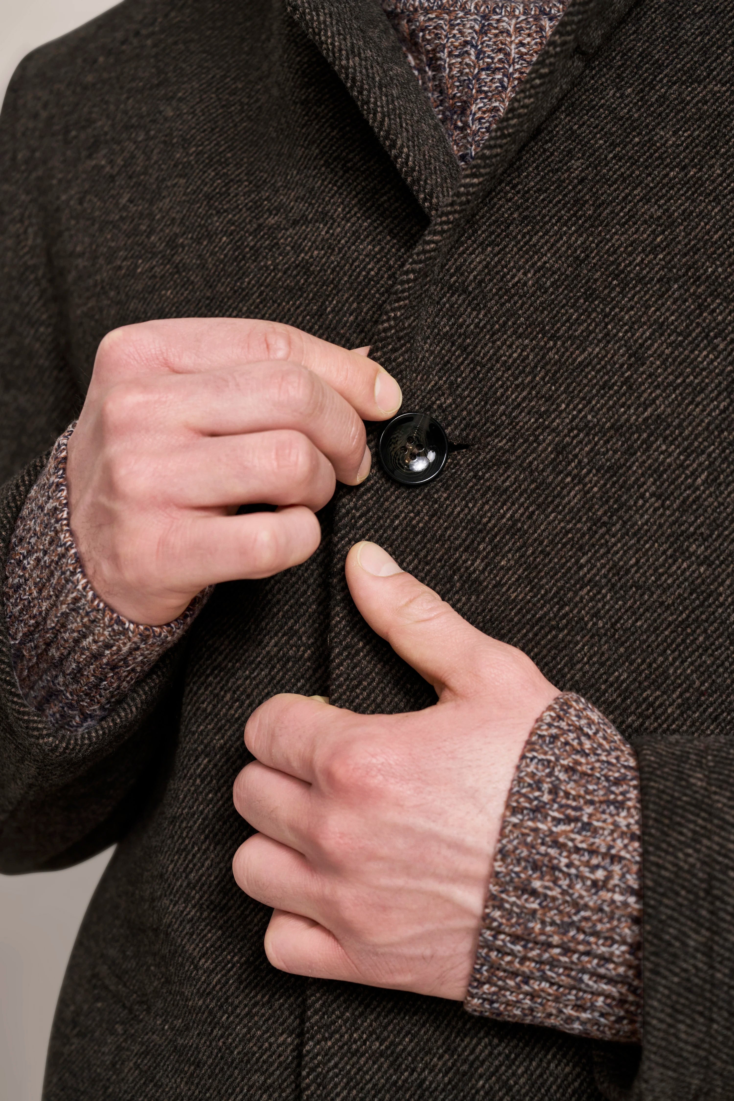 Herrenhand schließt Knopf an braunem bugatti-Mantel über Strickpullover. Büstenbild.