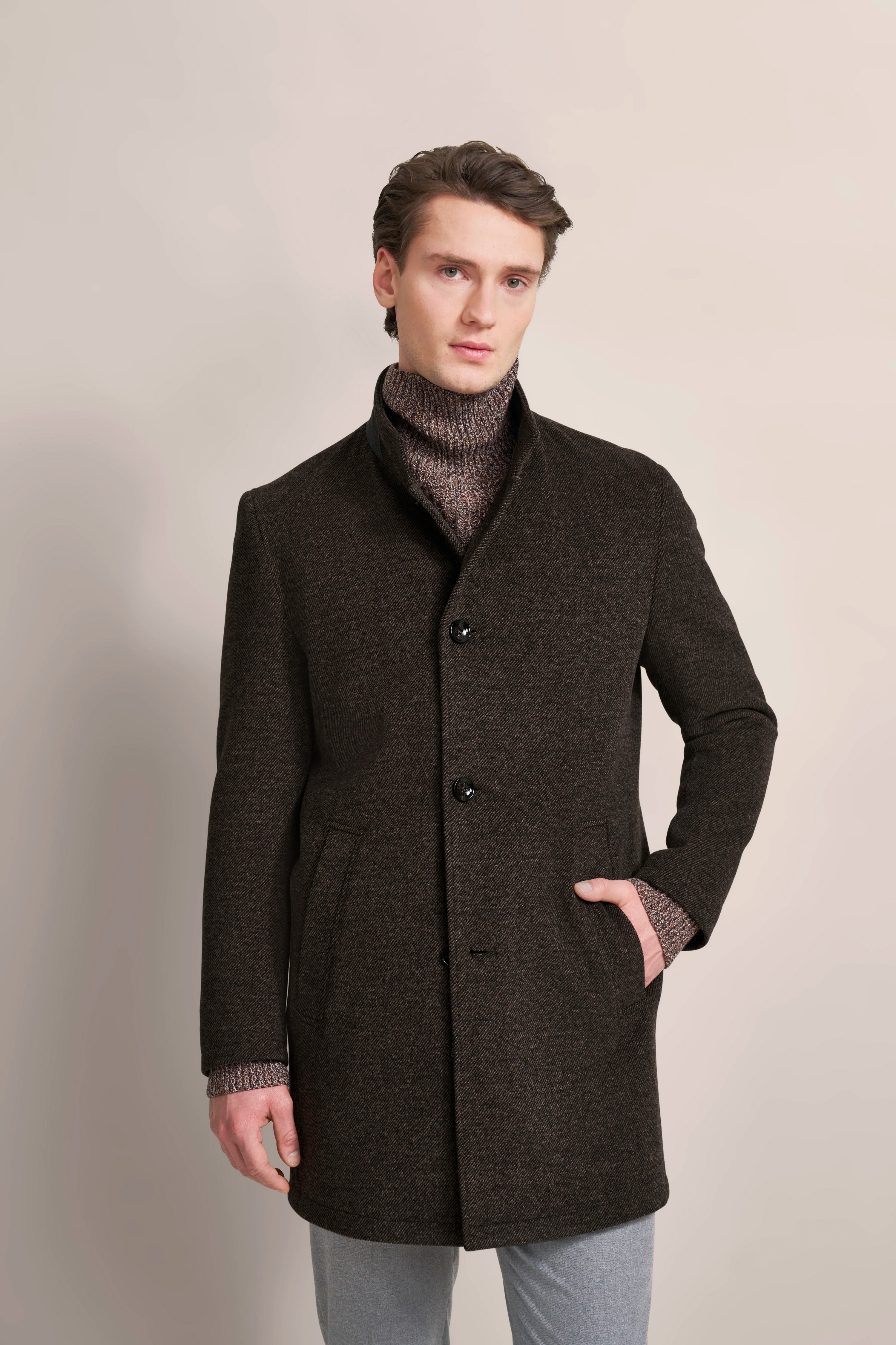 Model in brown bugatti coat, gray pants, posing with one hand in pocket.