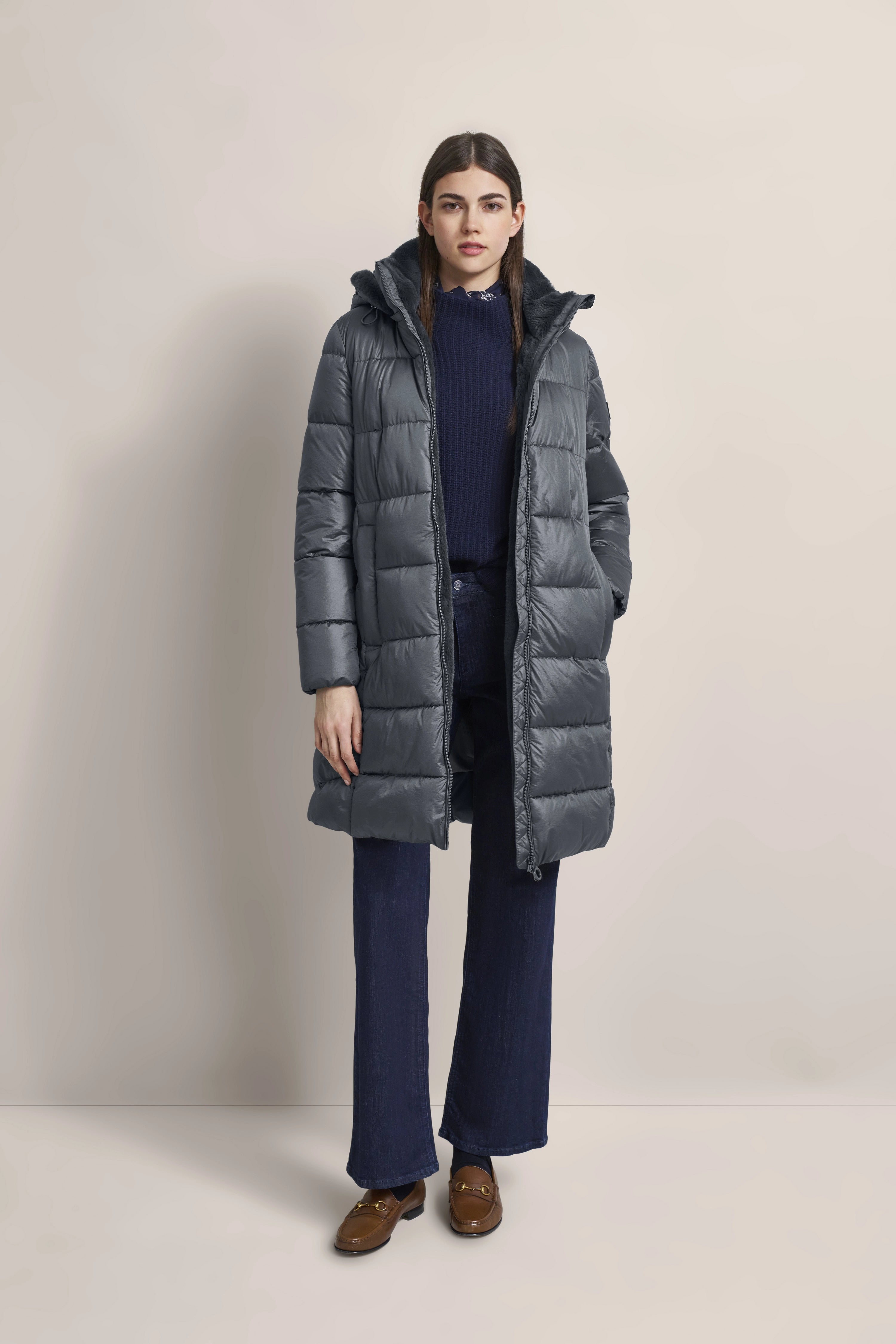 Model in grauem bugatti Daunenmantel, dunkelblauem Pullover, blauen Jeans, braunen Schuhen, Posing stehend.