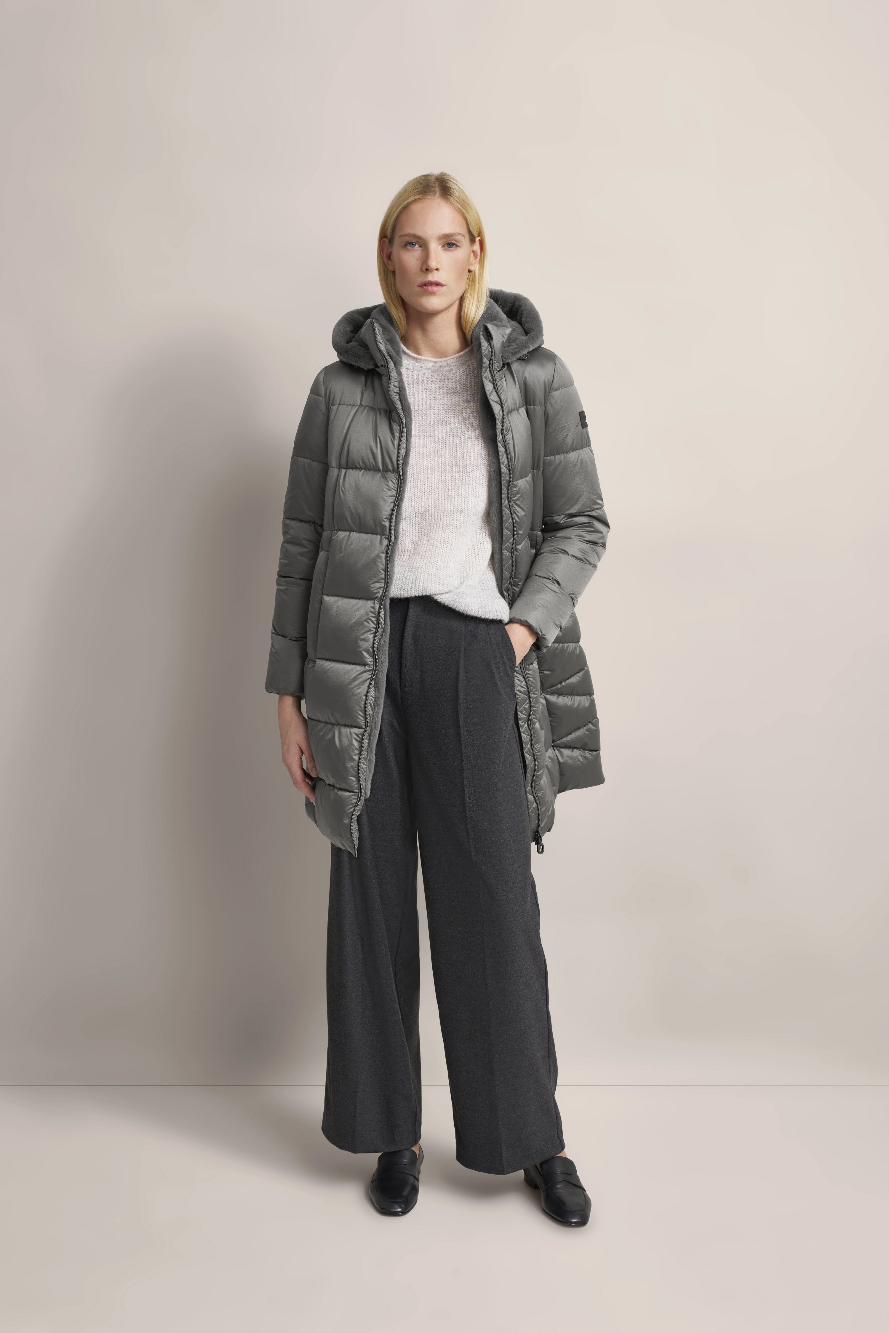 Grauer bugatti Daunenmantel, weißer Pullover, weite graue Hose, schwarzes Schuhwerk, Model stehend frontal.