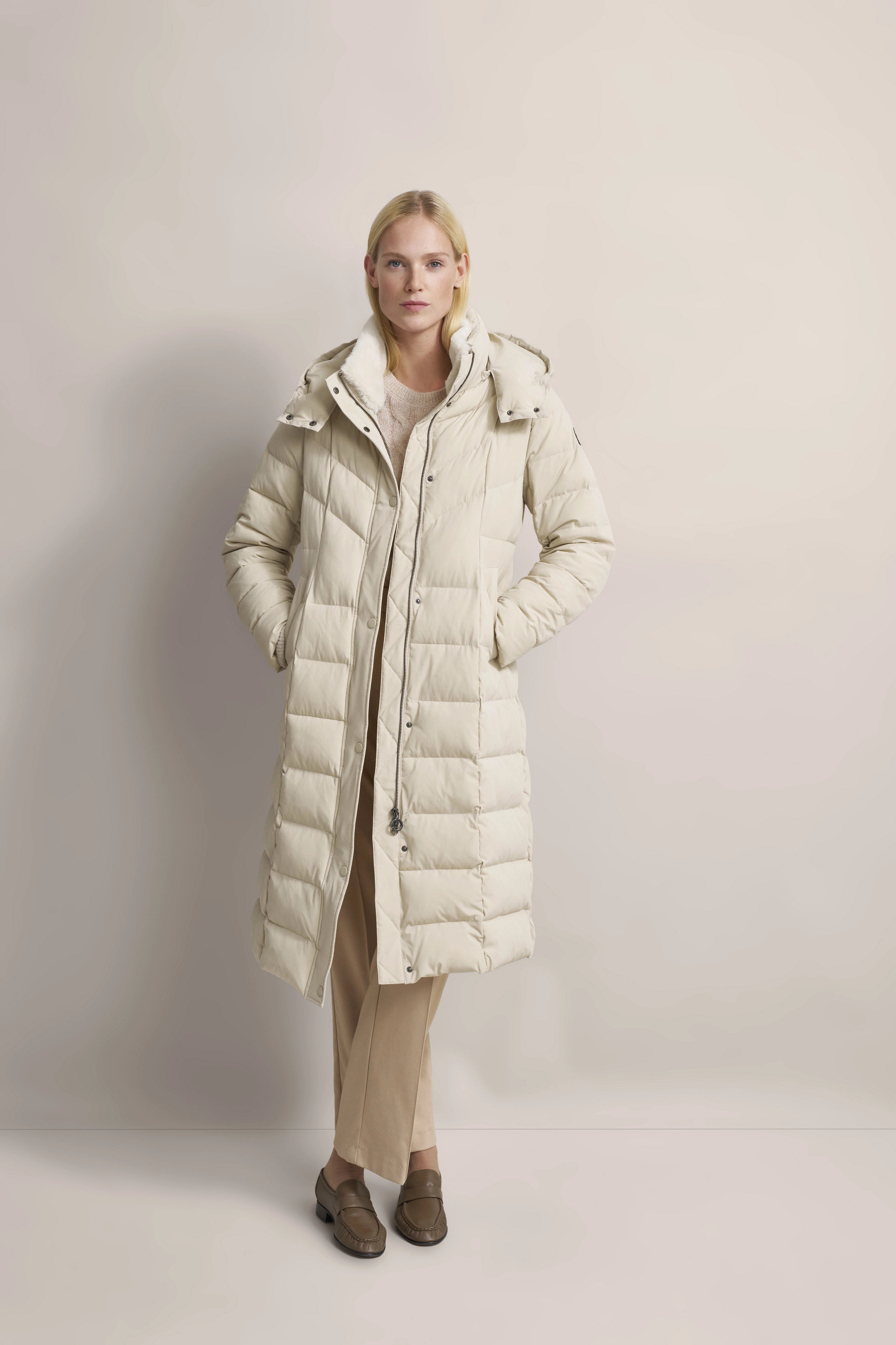 Model in cremefarbener bugatti Daunenjacke und beigen Hosen, lässige Pose, Hände in den Taschen.