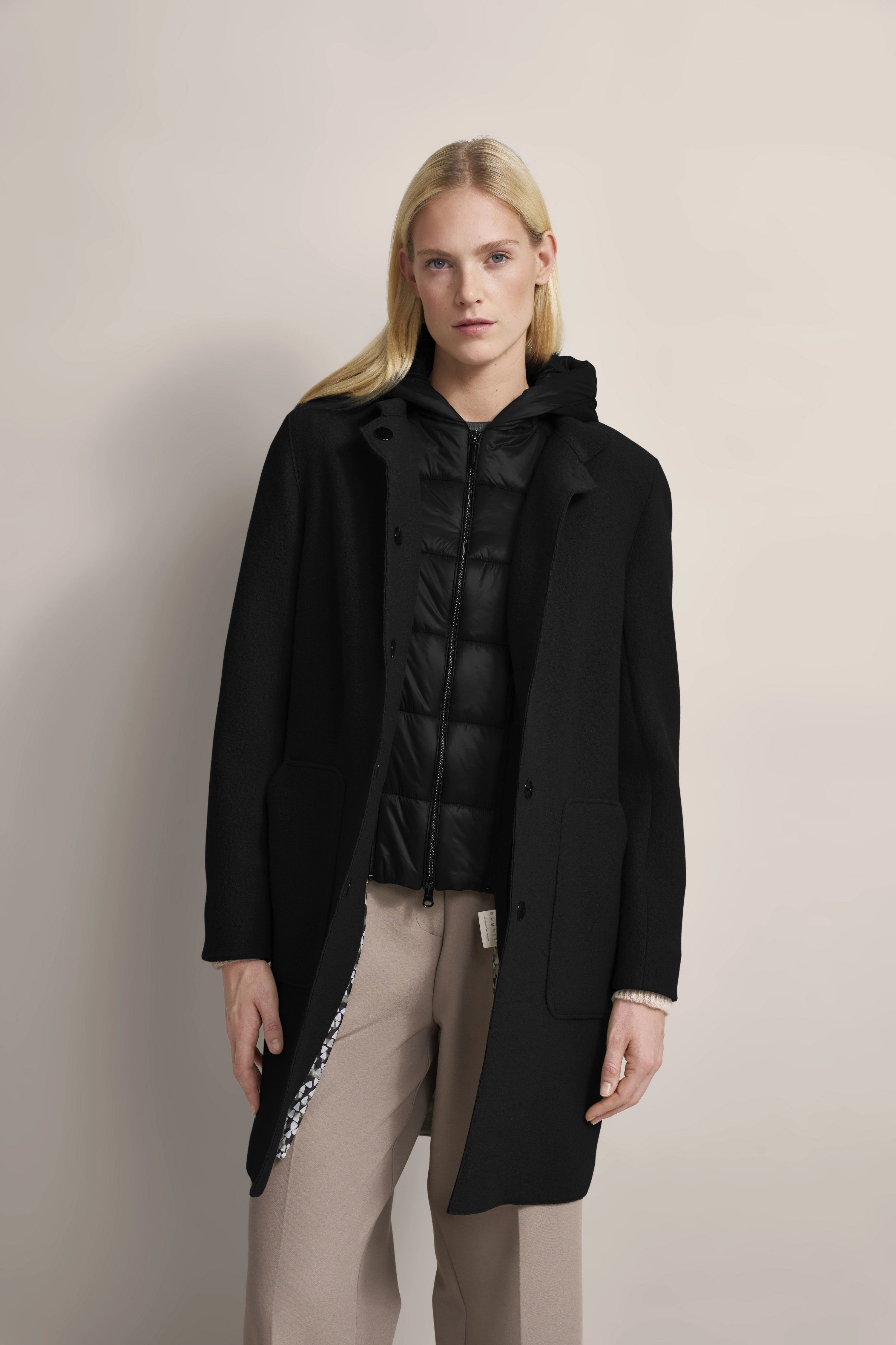 Elegant model in black bugatti coat, black quilted jacket, beige pants, relaxed pose.