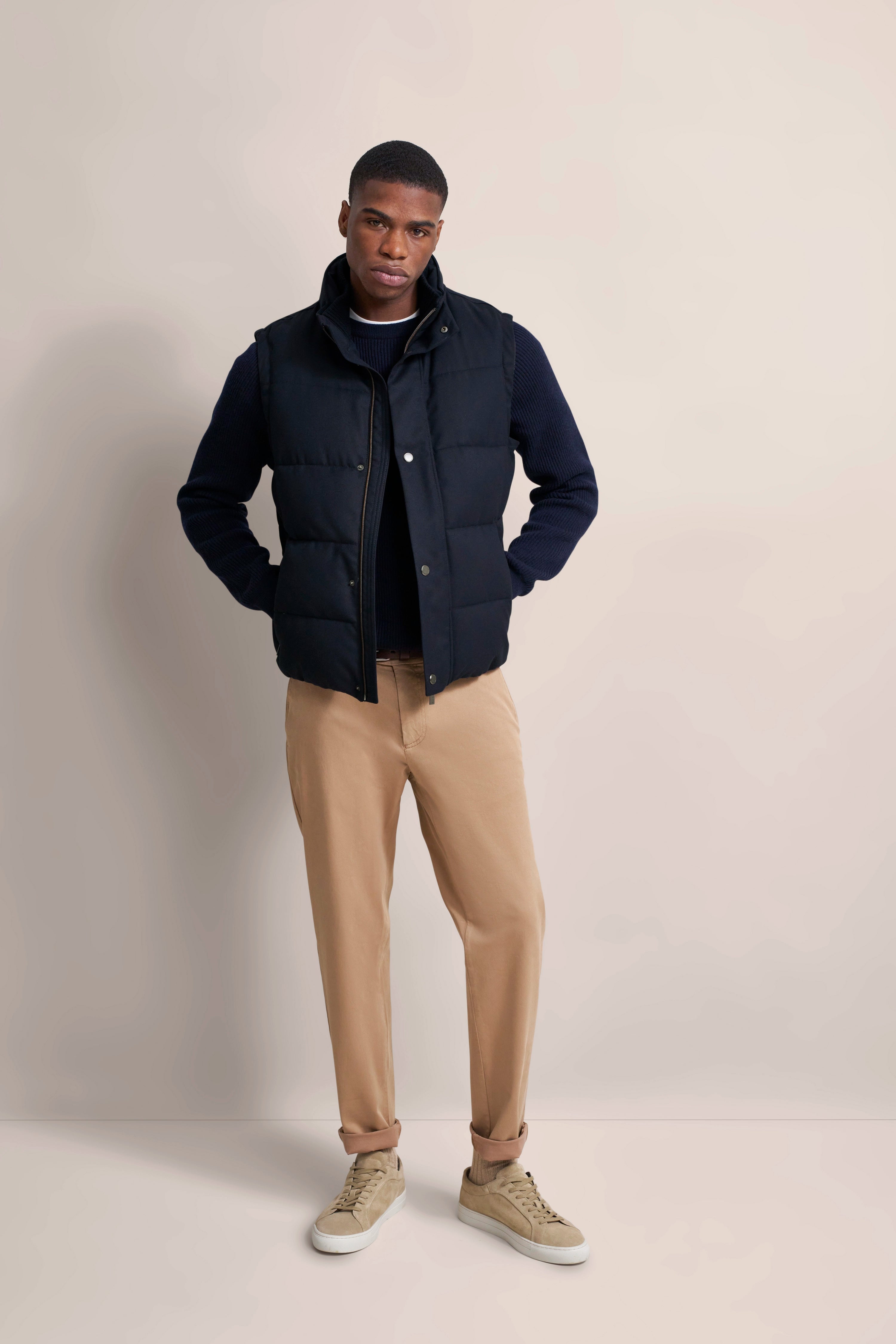 bugatti model shot: Black quilted vest, dark blue sweater, beige pants, beige sneakers, casually with hands in pockets.