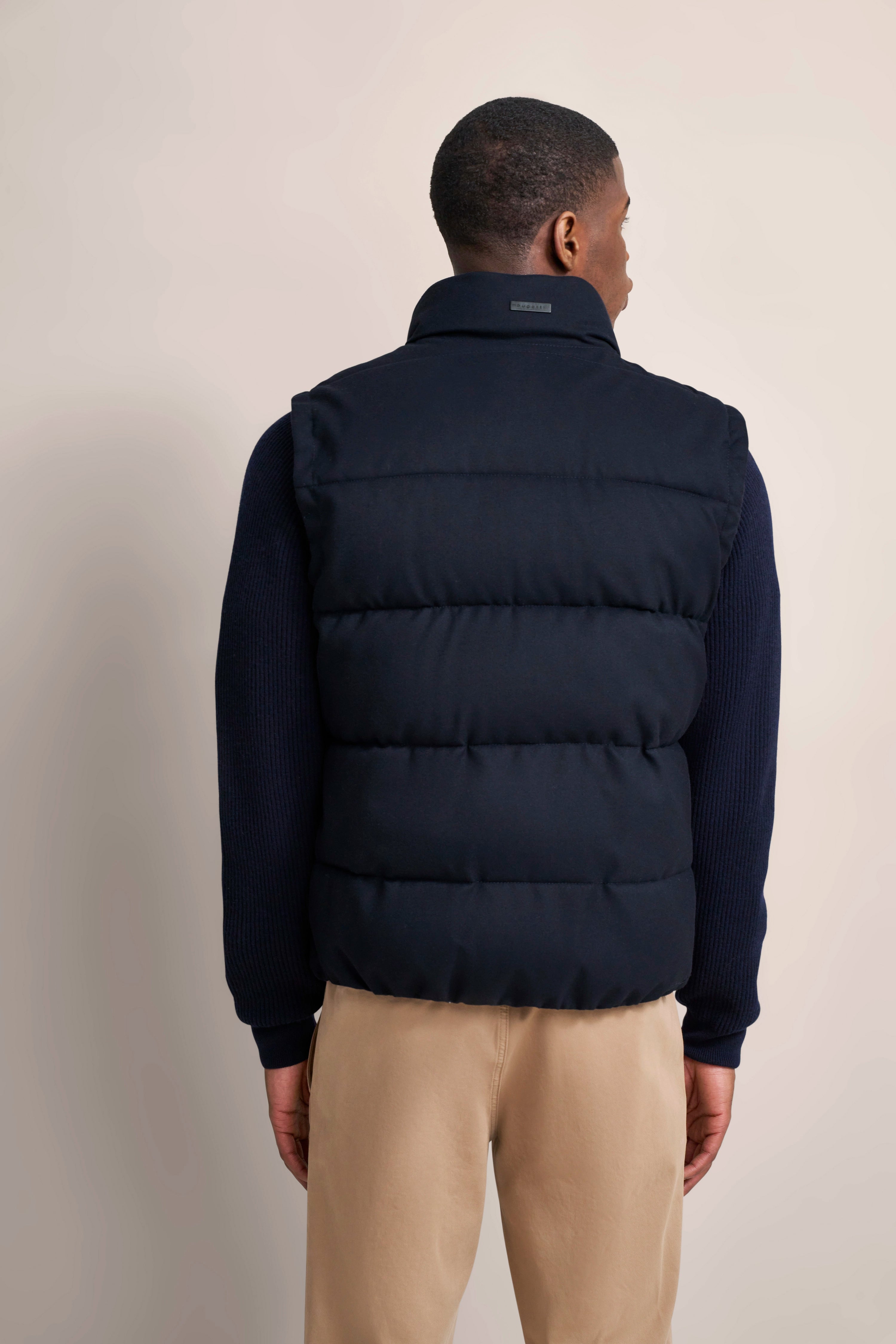 bugatti model: Navy vest, dark blue sweater, beige pants. Model from behind, relaxed posture.