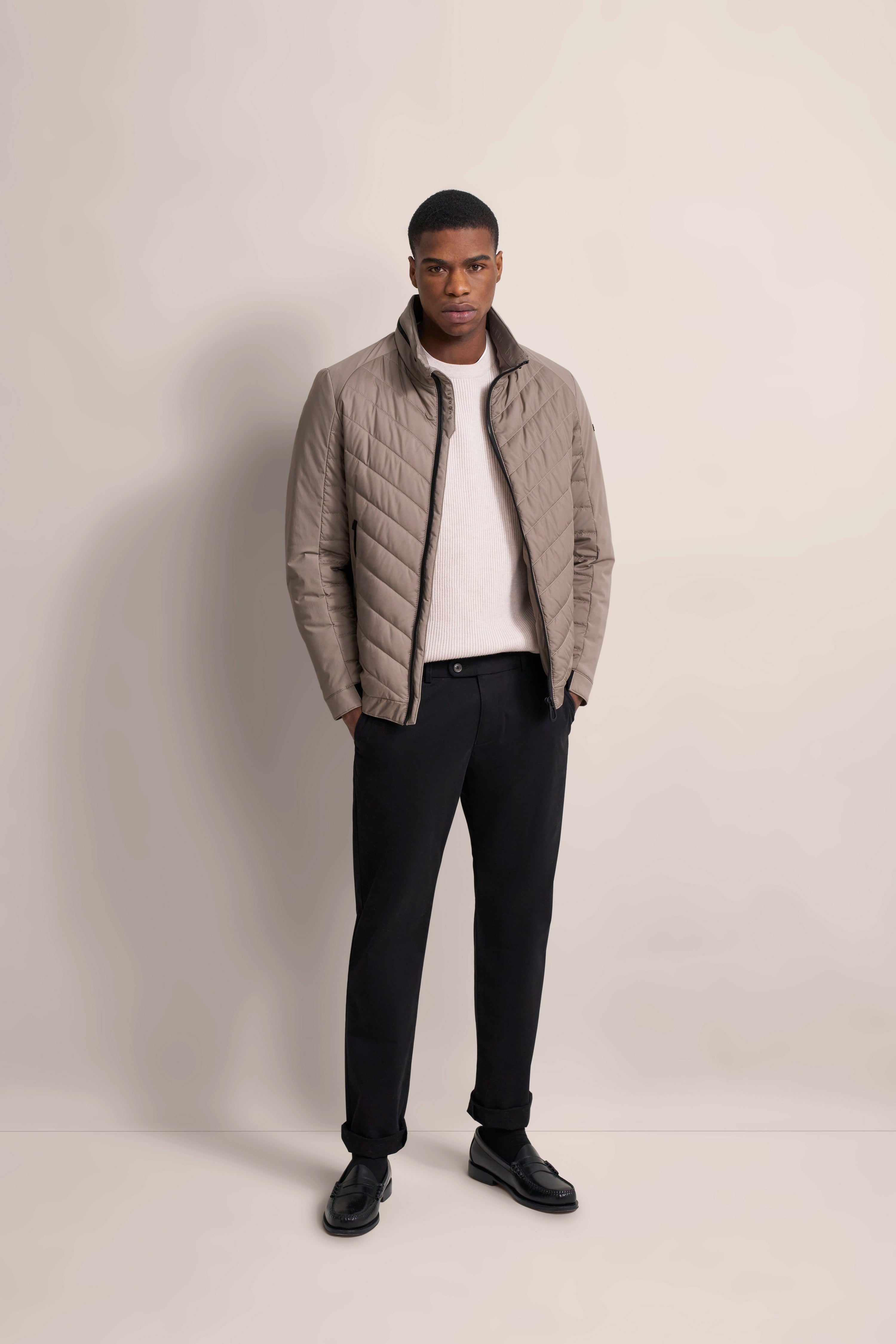 Model in beige quilted jacket, white sweater, black pants. Relaxed pose.