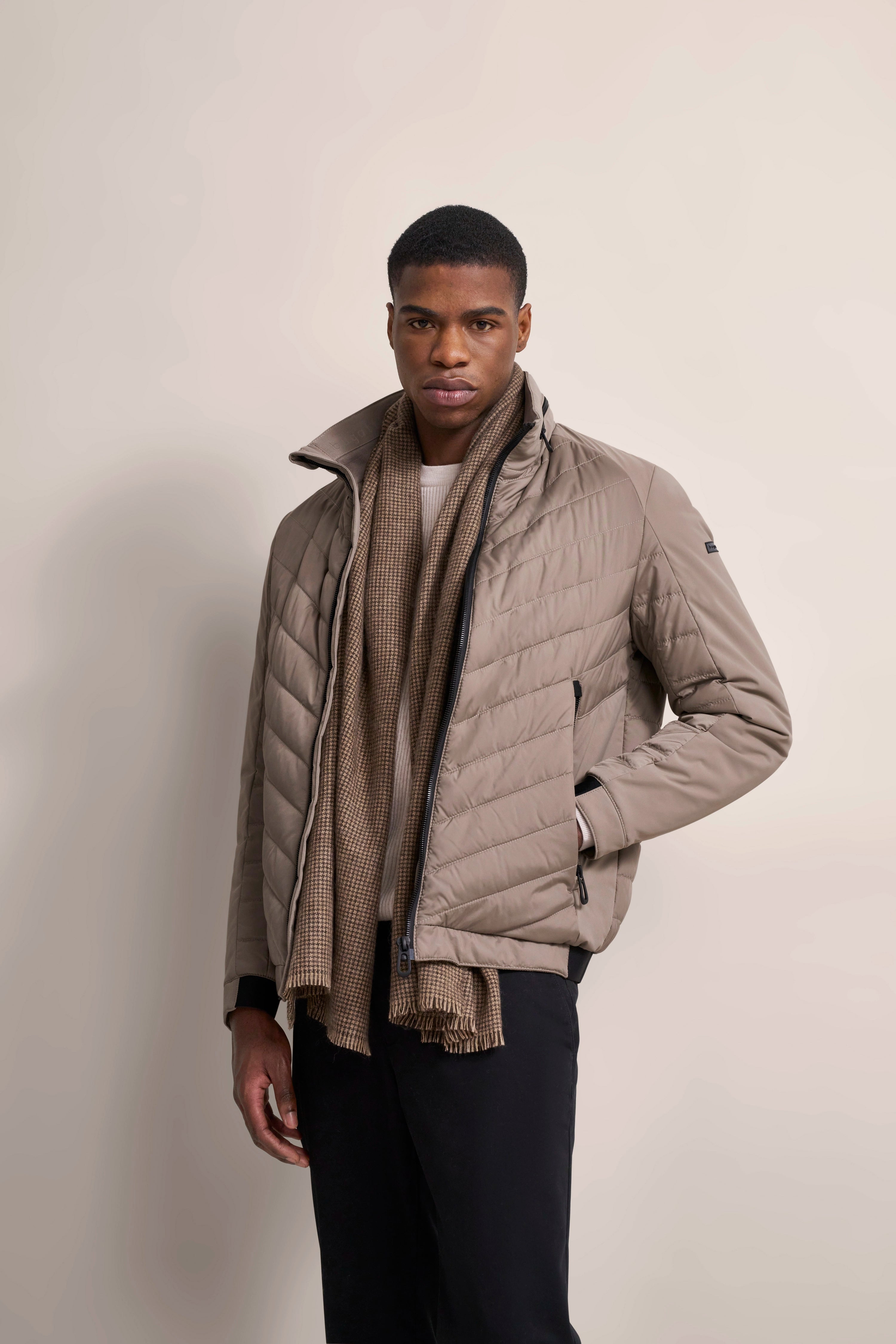 Model in beige quilted bugatti jacket, beige scarf, black pants, with a casual stance.