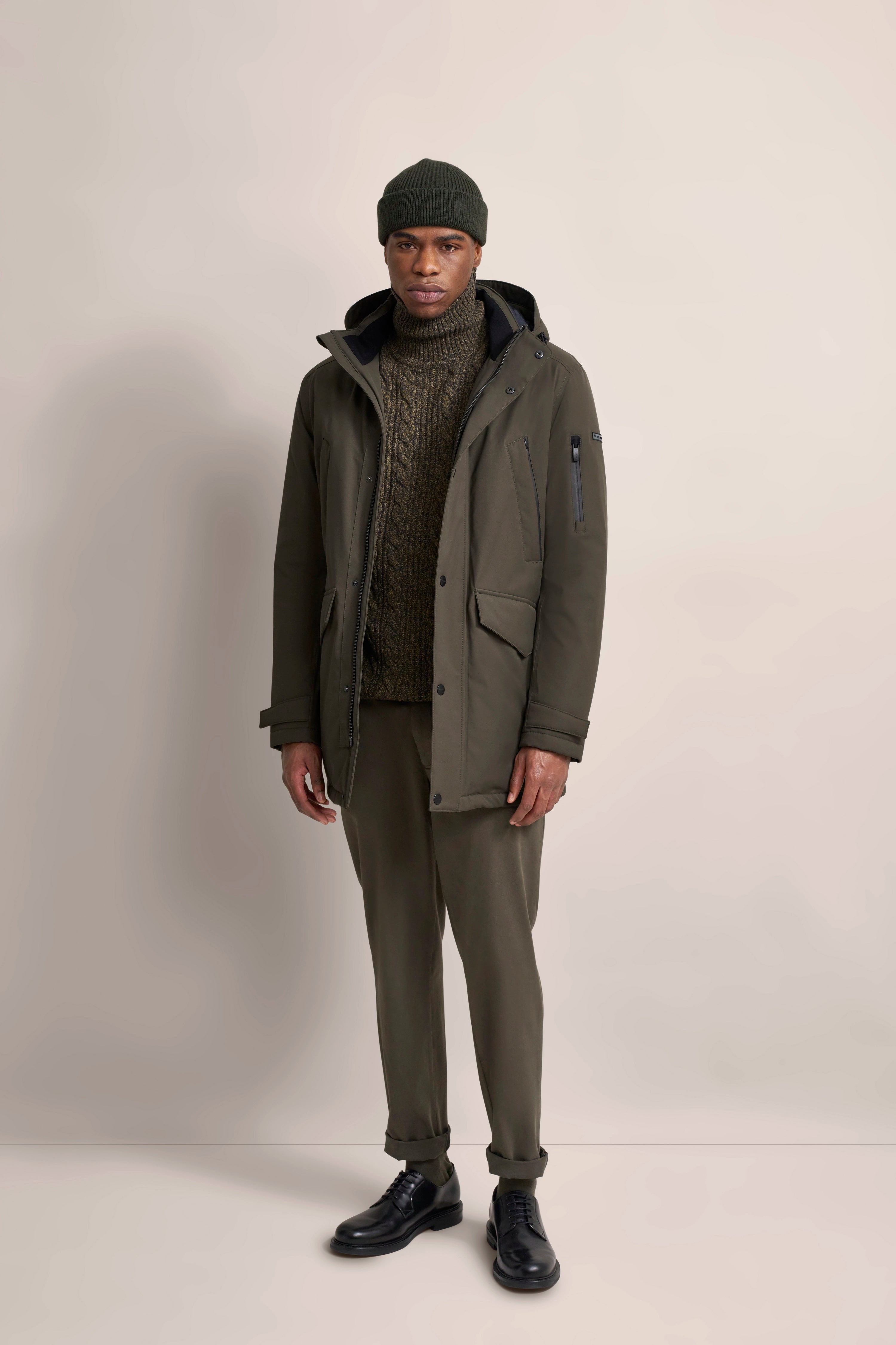 Model in olivgrünem Outfit mit Strickpullover, Parka, Hose und Mütze. Lässig stehend.