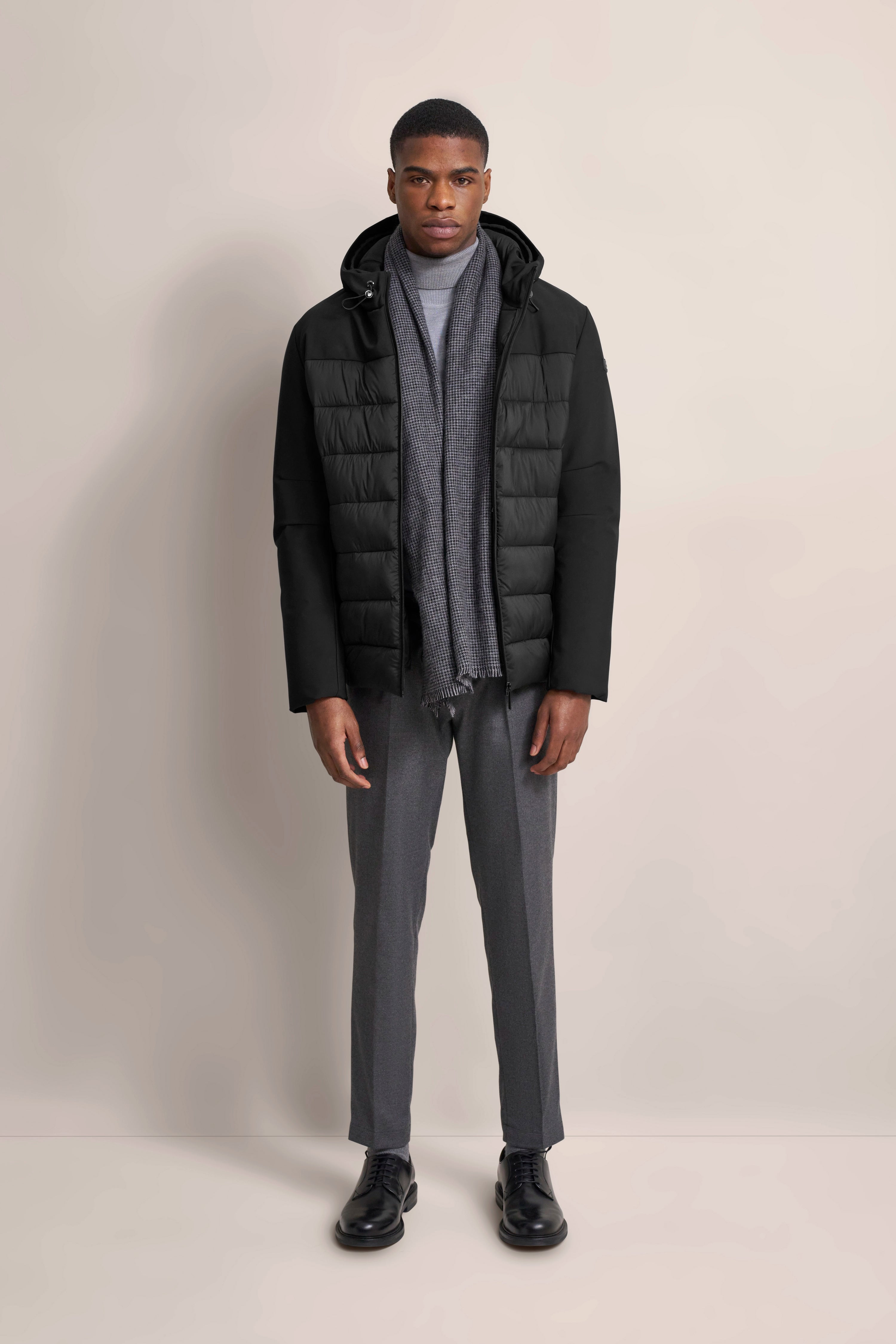 Model in schwarzer bugatti Jacke, grauem Schal und grauer Hose. Posing: gerade stehend, frontal.