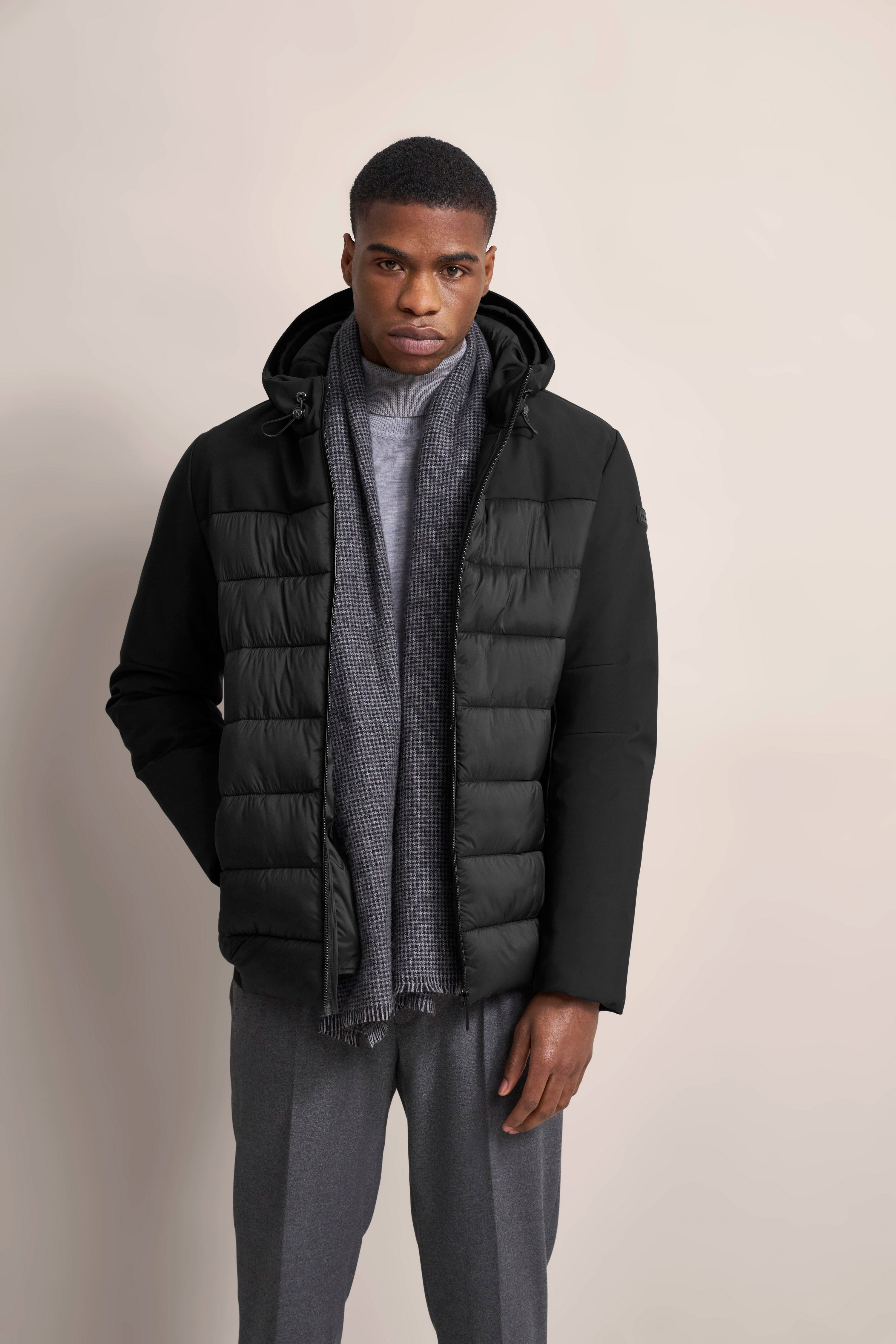 Model in schwarzer bugatti Jacke, grauem Schal und grauer Hose, frontal mit Händen in den Taschen.