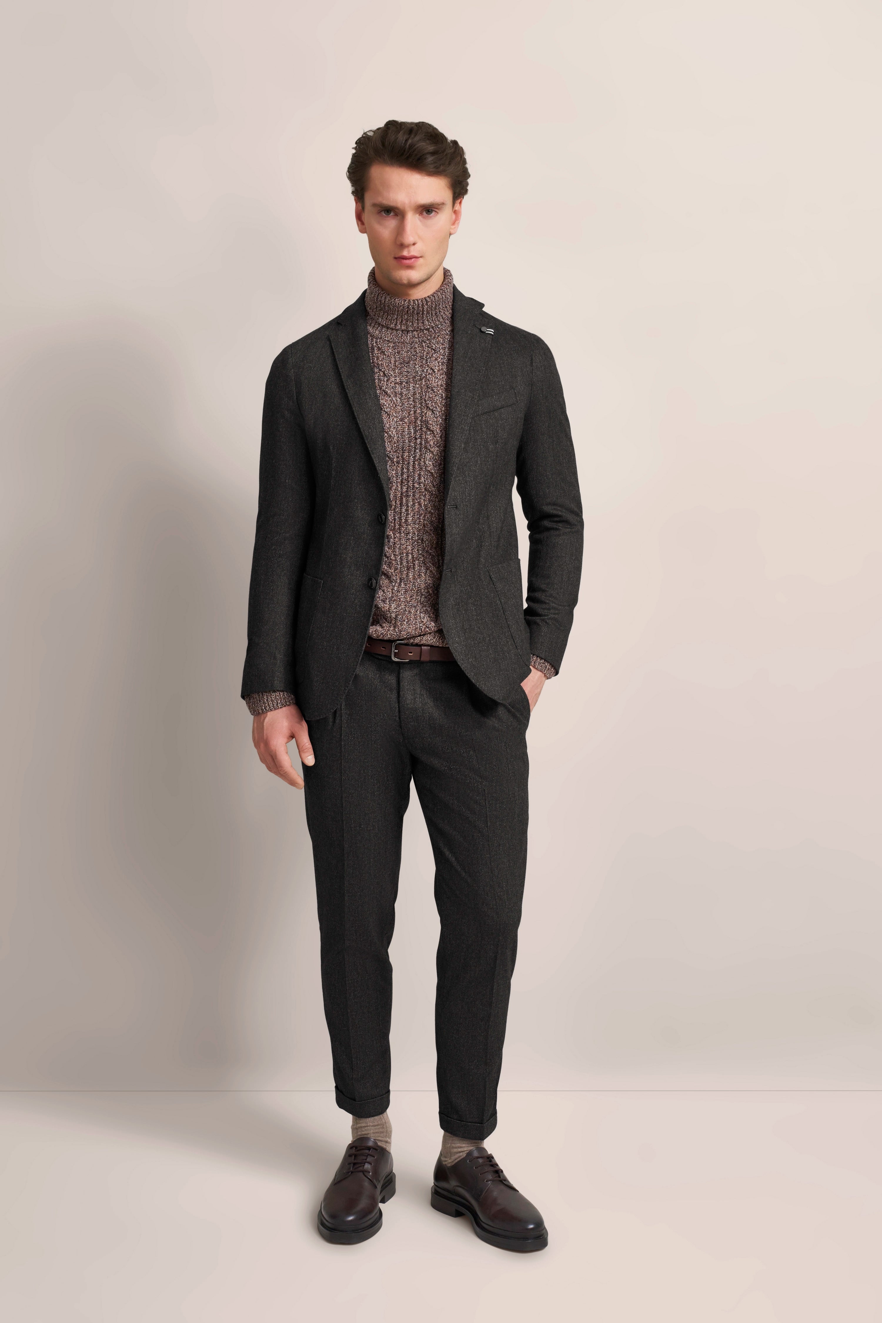 Model in dunklem Anzug mit grauem Rollkragenpullover, stehend, Hände in Taschen.