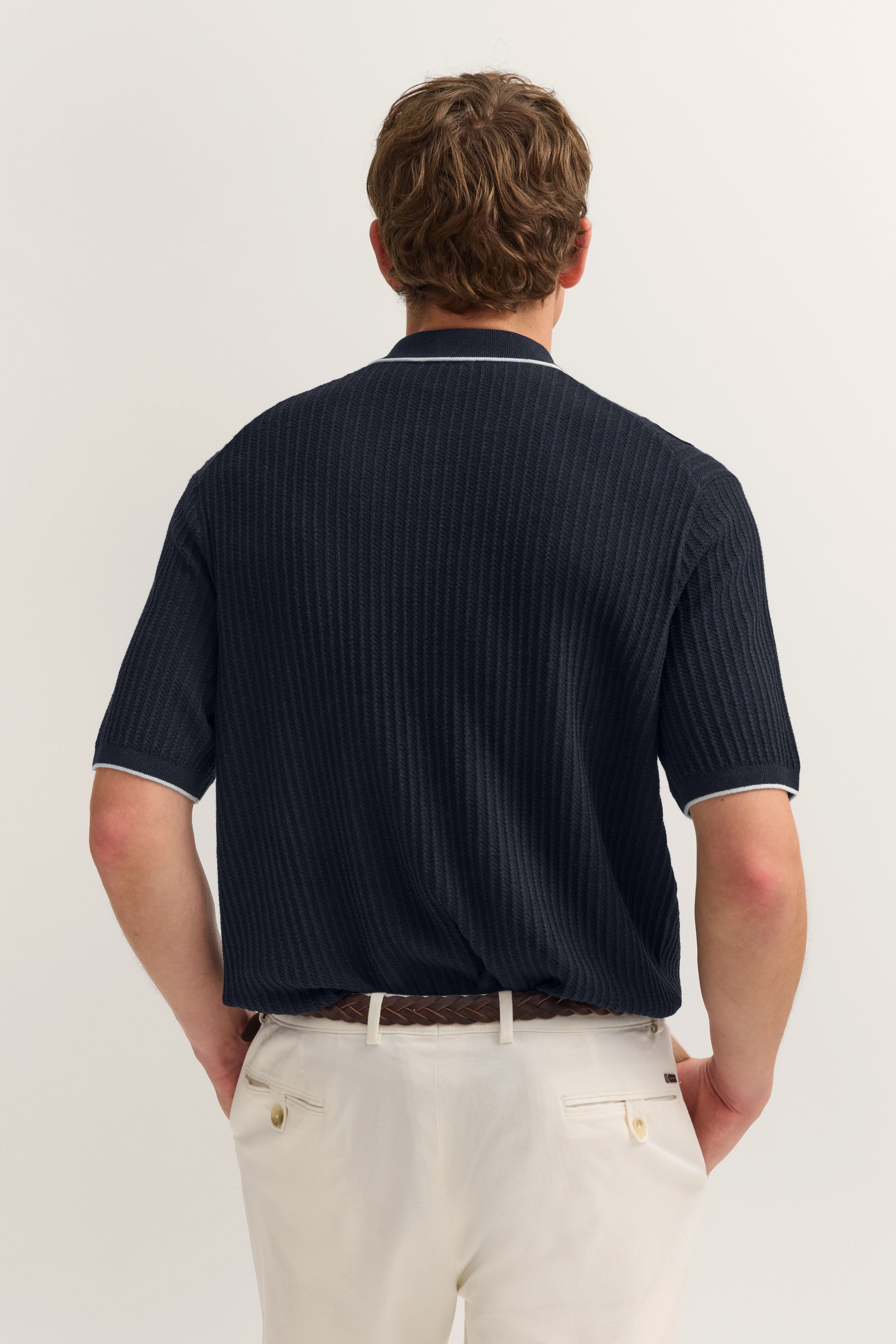 bugatti Model von hinten, trägt dunkelblaues Polo mit Struktur und cremeweiße Hose, legeres Posing.
