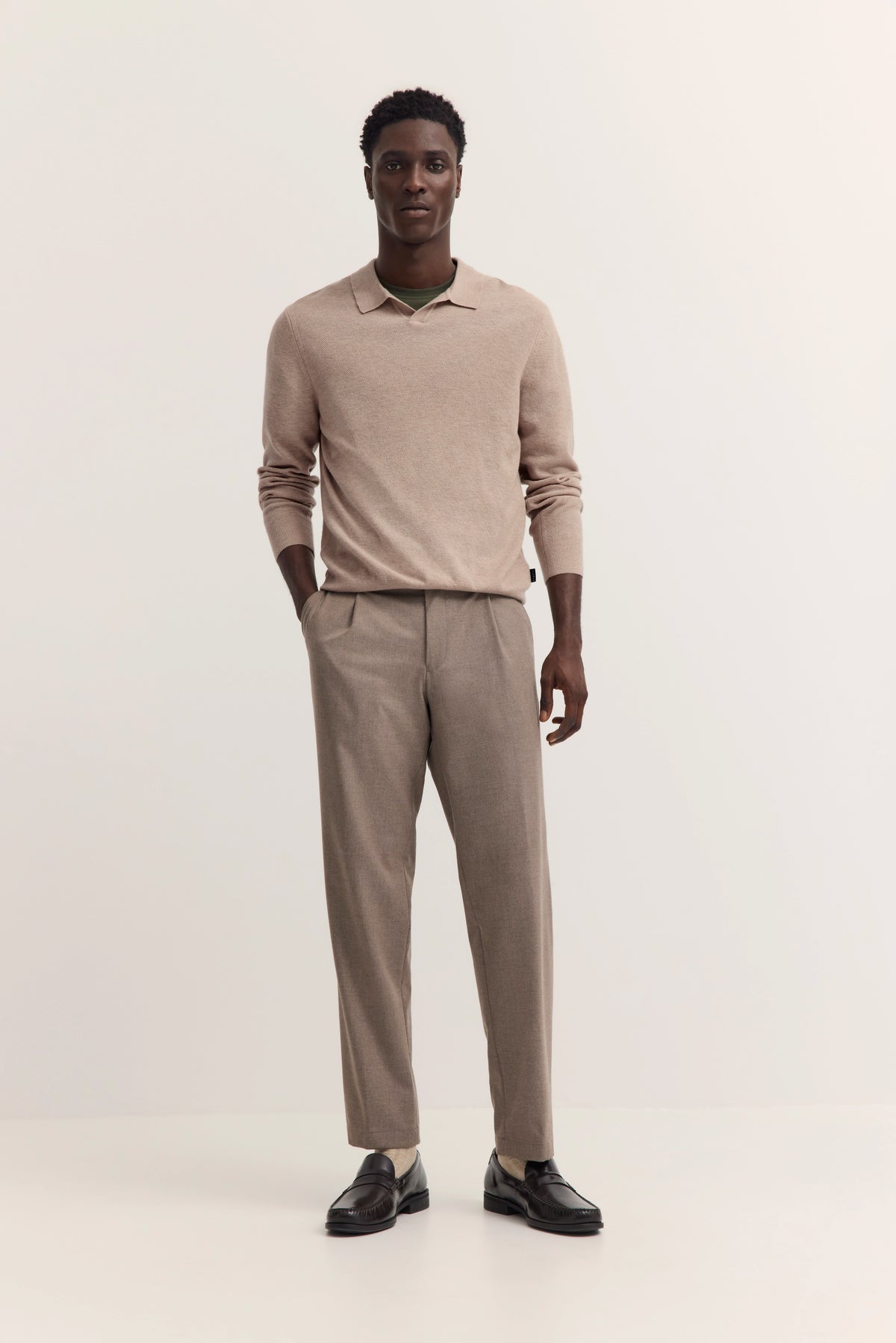 Model in beigem Pullover und Hose, schwarze Schuhe, entspannte Haltung. Bugatti Stil.