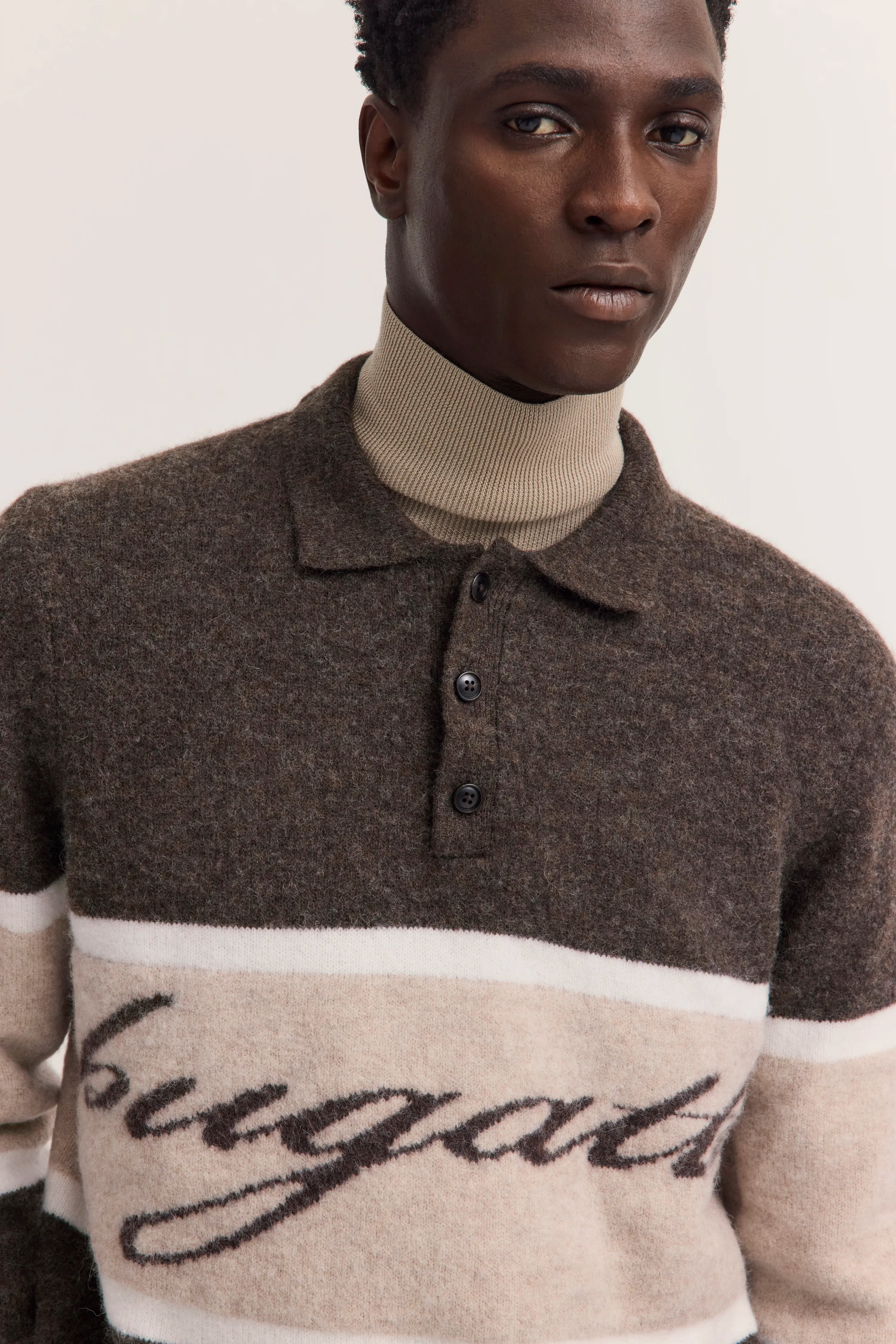 Büste von Mann im braunen bugatti-Pullover mit Schriftzug und beigem Rollkragen.
