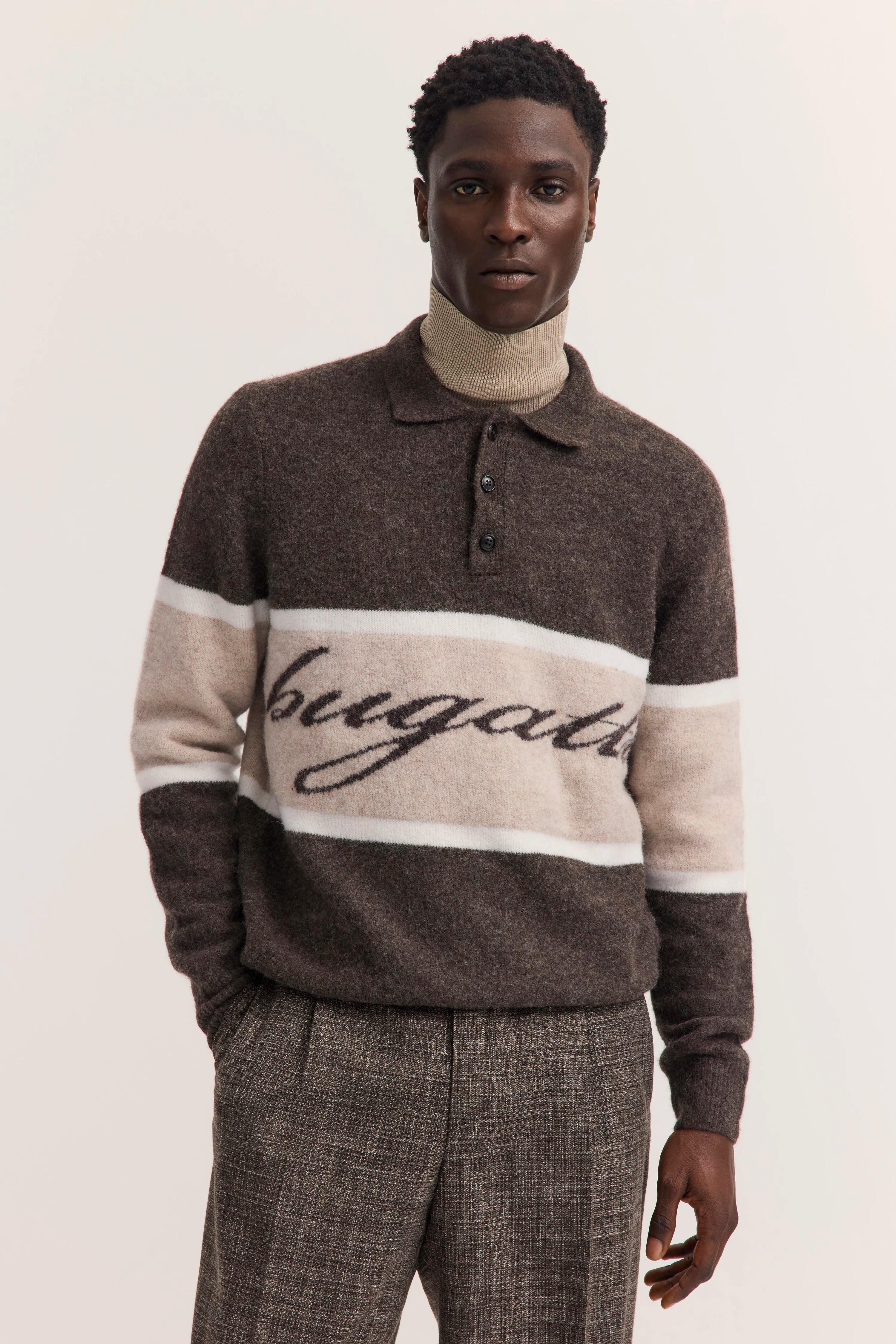 Brauner bugatti Pullover mit Logo, beige Rollkragen, graue Hose. Modelbild frontal, entspannte Haltung.