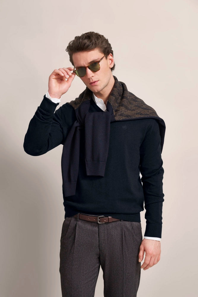 Model in dark blue sweater, gray pants, scarf over shoulder, sunglasses, casual pose.