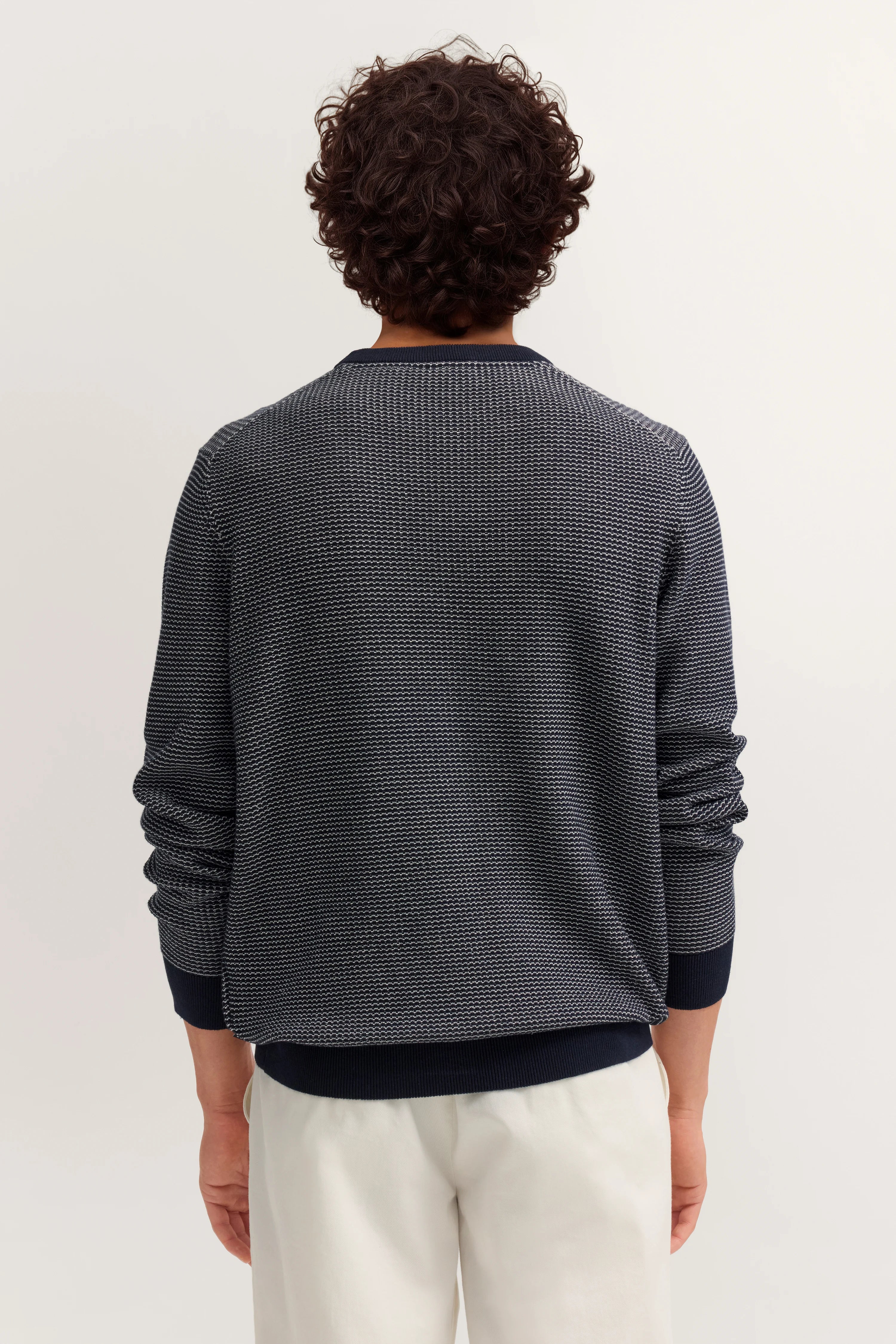 Rückansicht eines Models in dunkelblauem bugatti Pullover, legeres Outfit mit weißen Hosen.