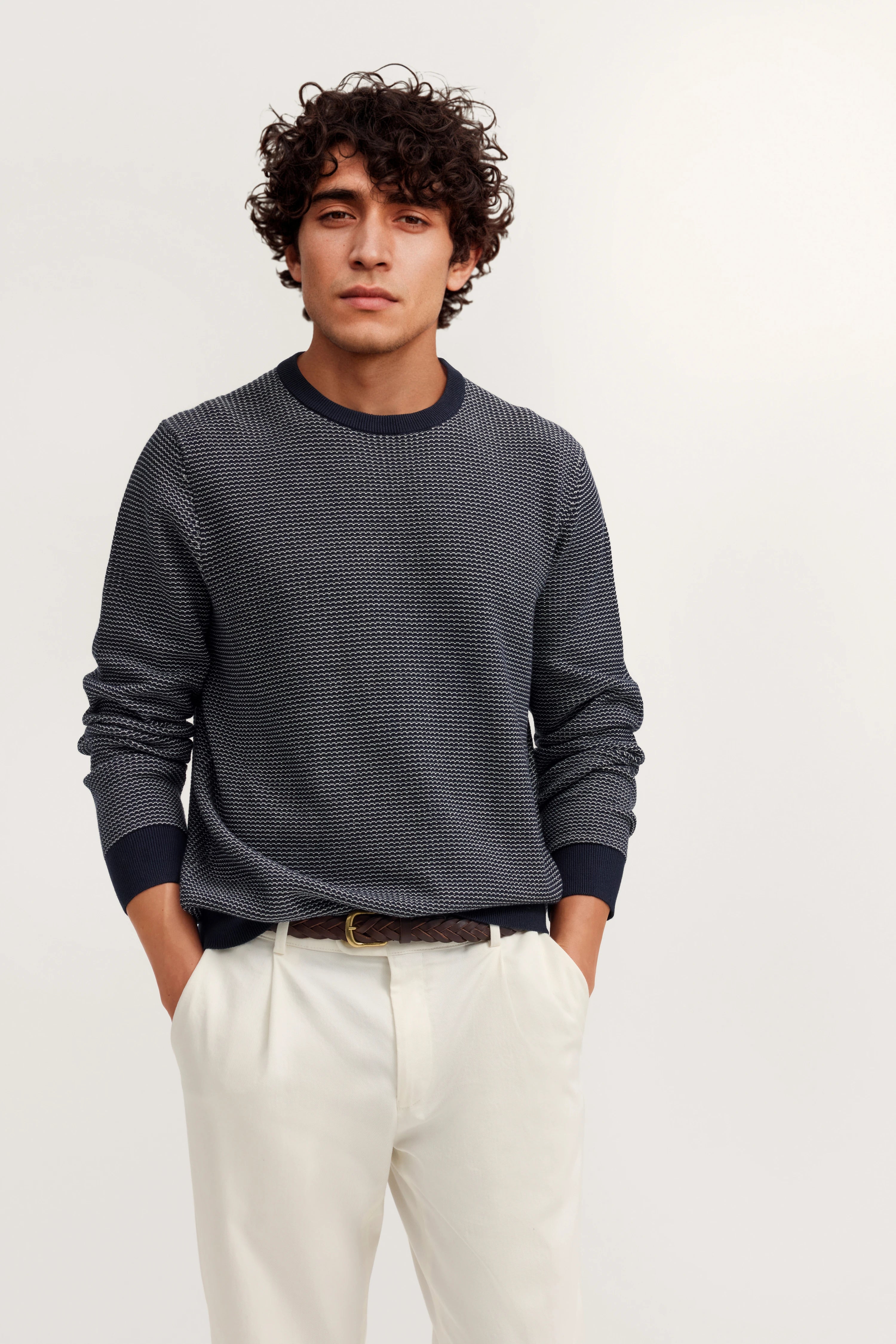 Model in navy gemustertem Pullover und weißen Hosen, lässige Pose, Hände in den Taschen.