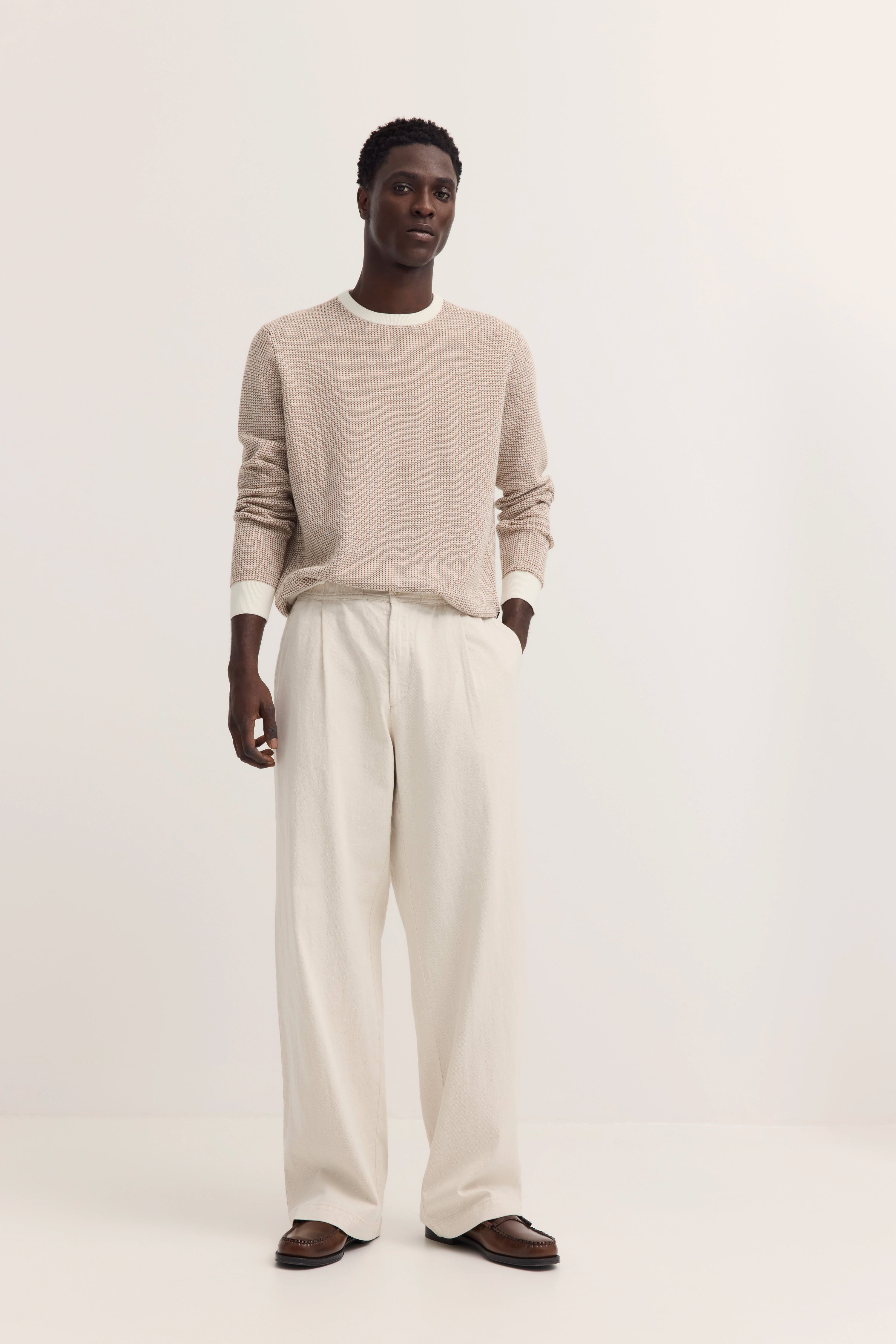 Beiger Strickpullover mit weißen Bündchen, weite cremefarbene Hose, braune Schuhe, Model stehend.