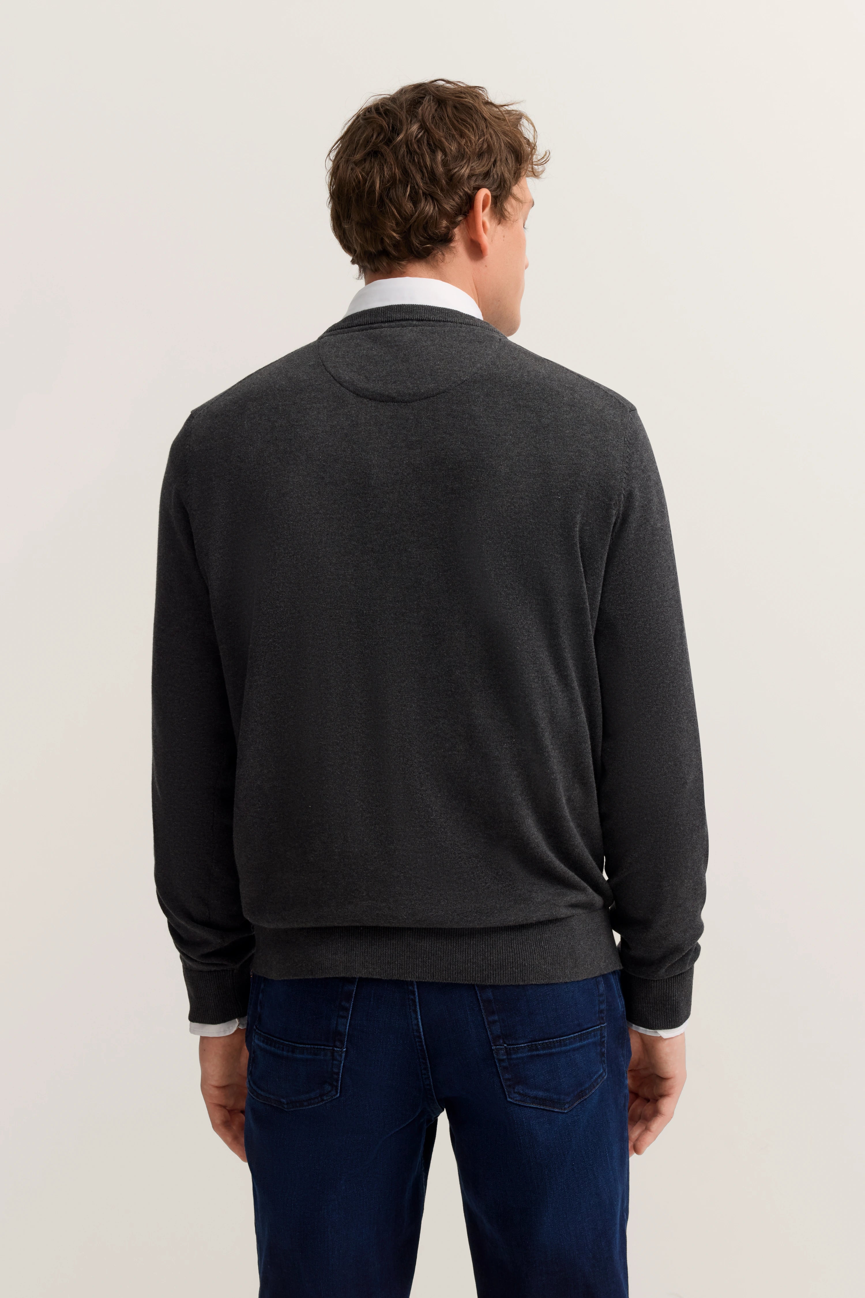 bugatti Herren-Outfit: Dunkler Pullover, weiße Hemdkante, blaue Jeans, Rückenansicht, legere Pose.