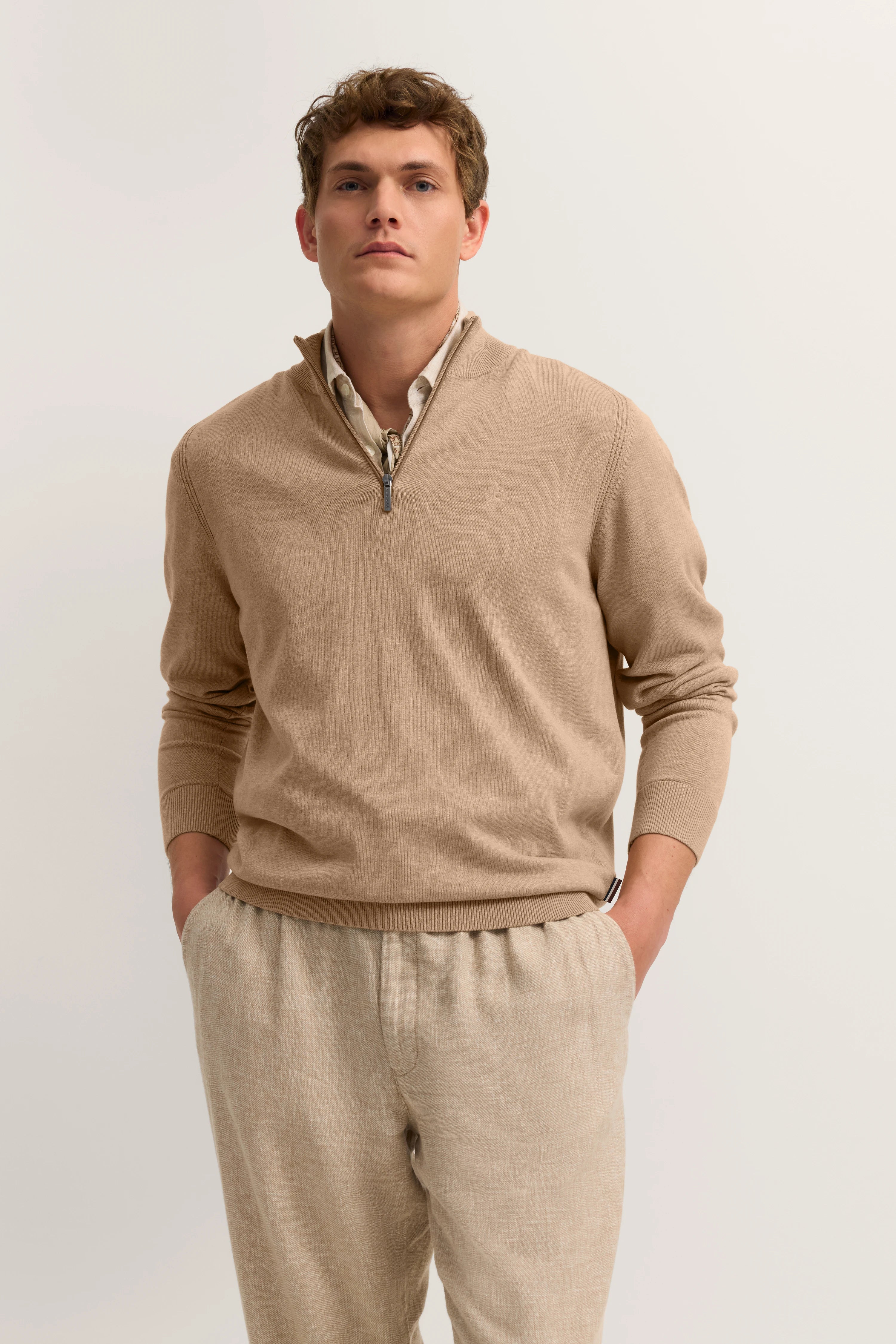 Beiger Pullover mit Reißverschluss, beige Hose, Model mit Händen in Taschen, entspanntes Posing.