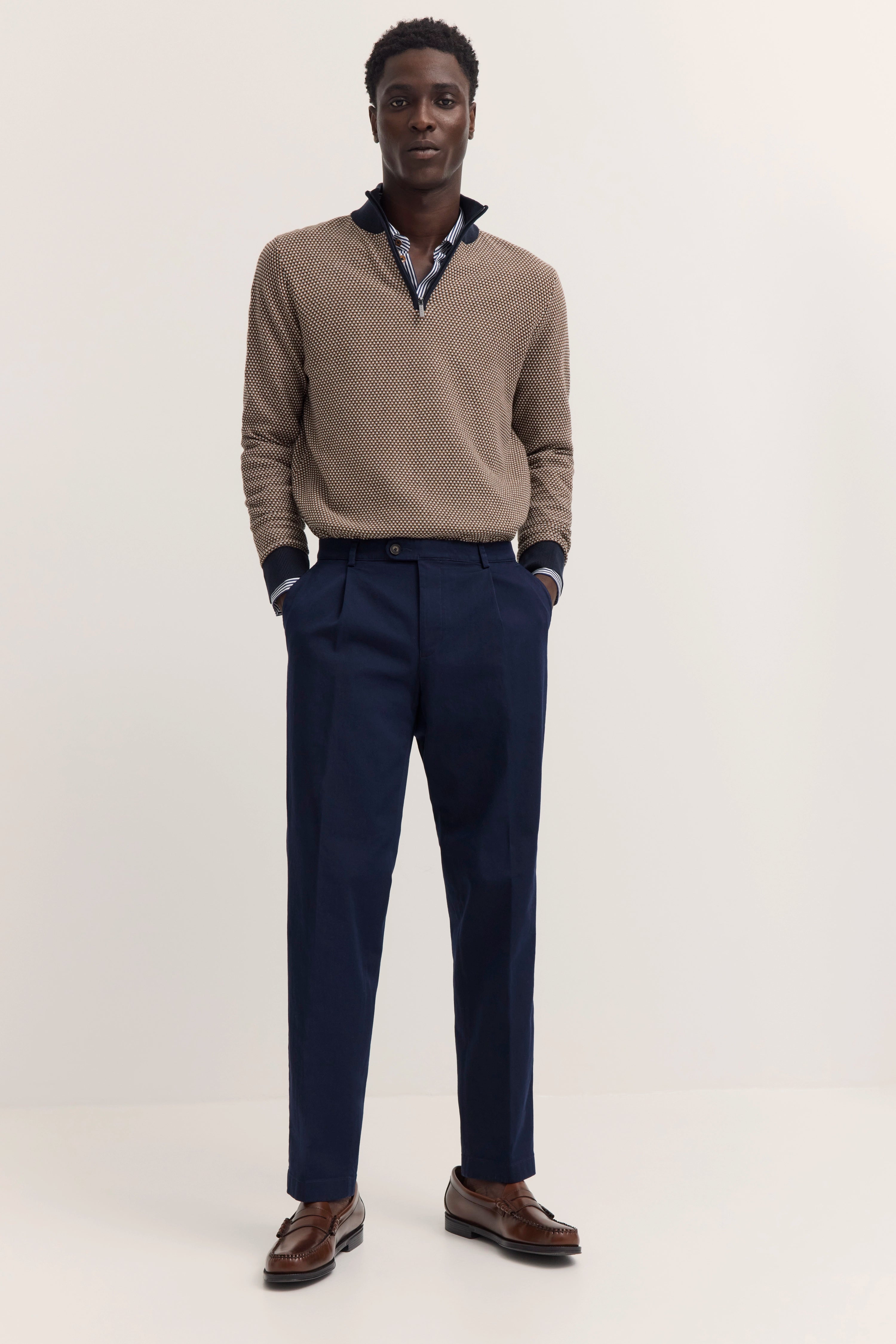 Model in beigem Pullover mit Muster, blauer Hose, braunen Schuhen, legeres Posing.