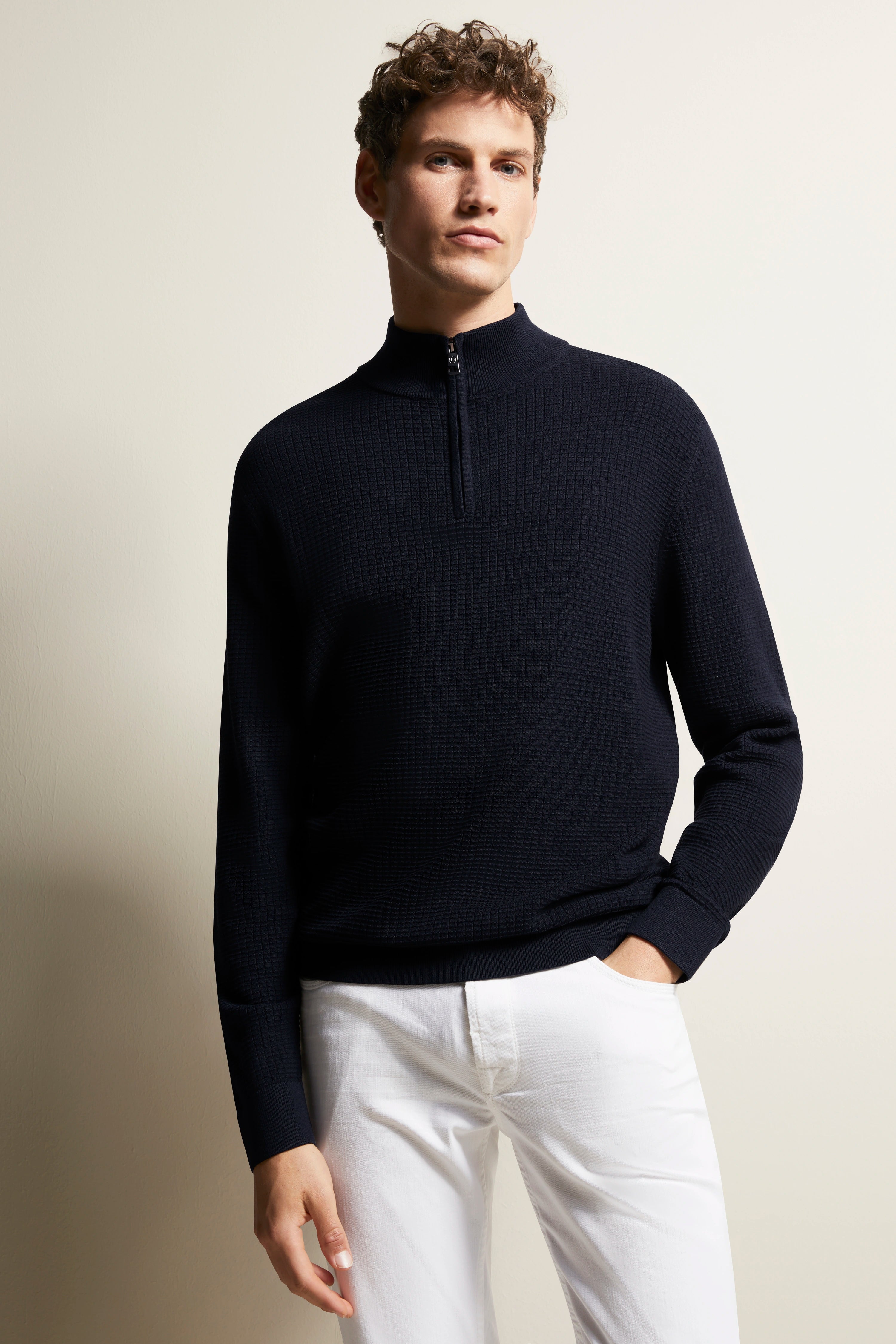 Model in navy bugatti zip sweater and white trousers, relaxed pose.