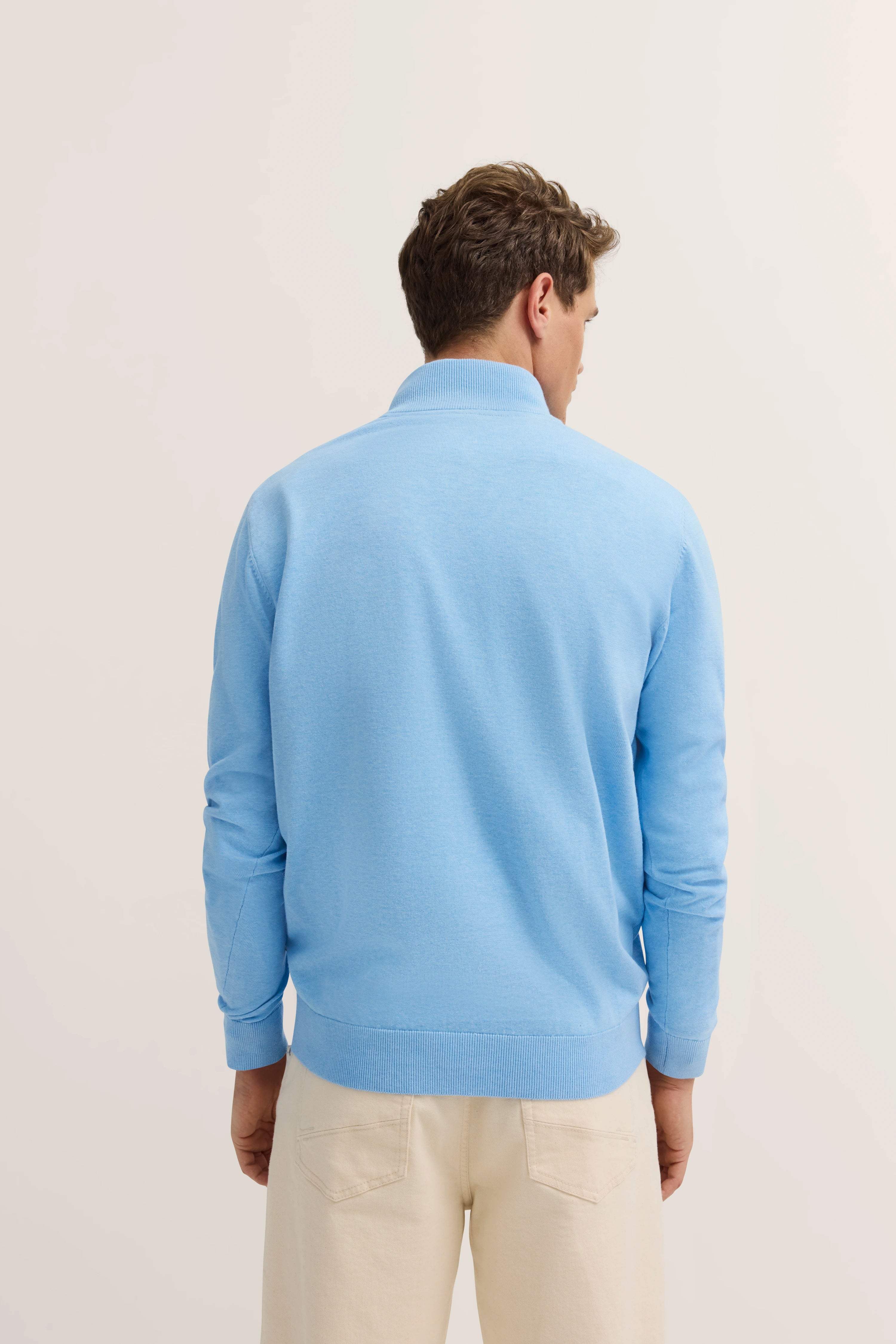 Model in hellblauem bugatti Pullover, Rückenansicht, cremefarbene Hose, legere Pose.