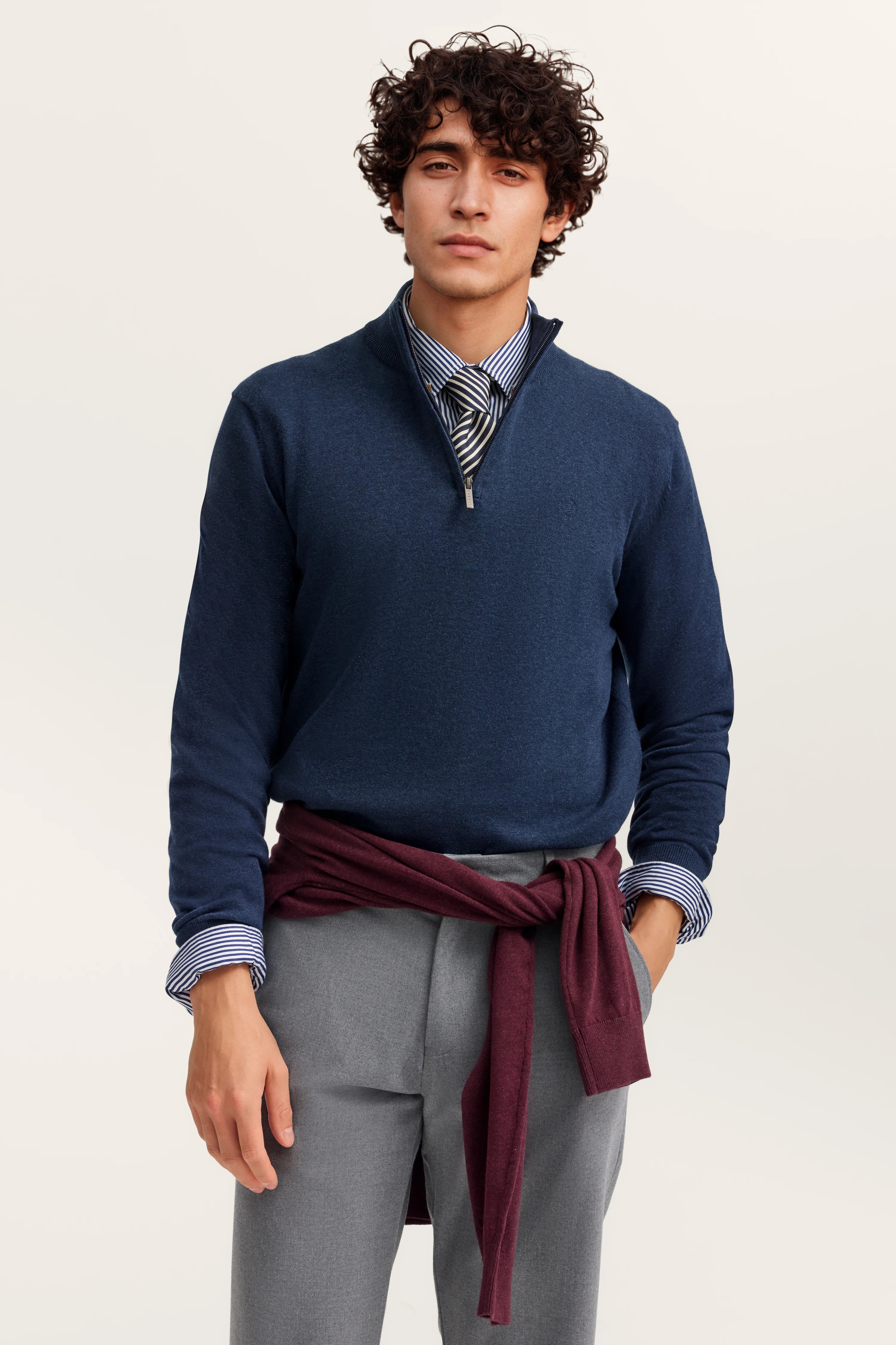 Model in blauem Pullover, gestreiftem Hemd, grauer Hose und bordeauxrotem Pullover um die Taille.