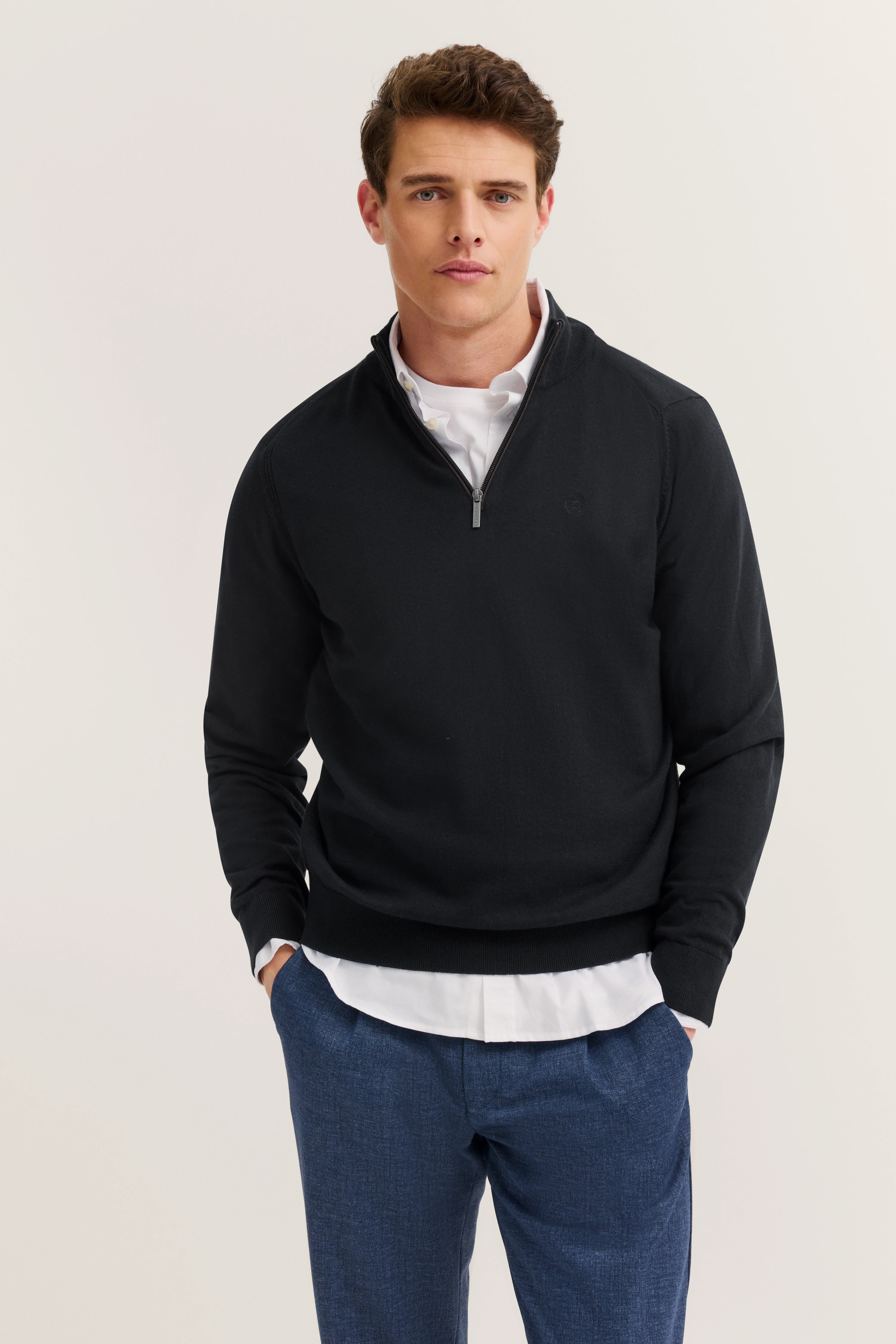 Model wears black bugatti zipper sweater, white shirt, blue pants. Hands in pockets.