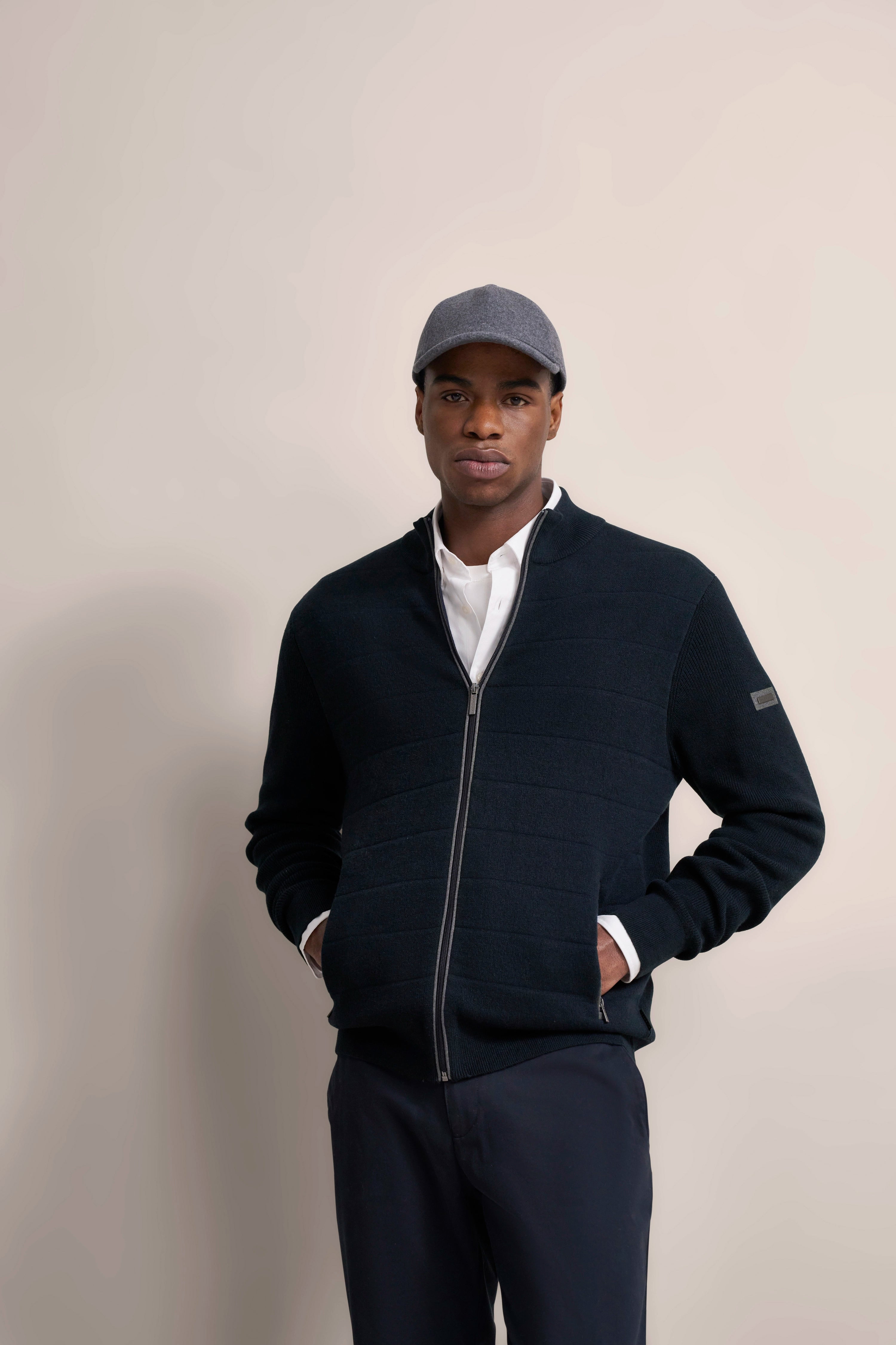 Model in dunkelblauer bugatti Jacke, weißem Hemd, grauer Mütze, legeres Posing, Hände in Taschen.