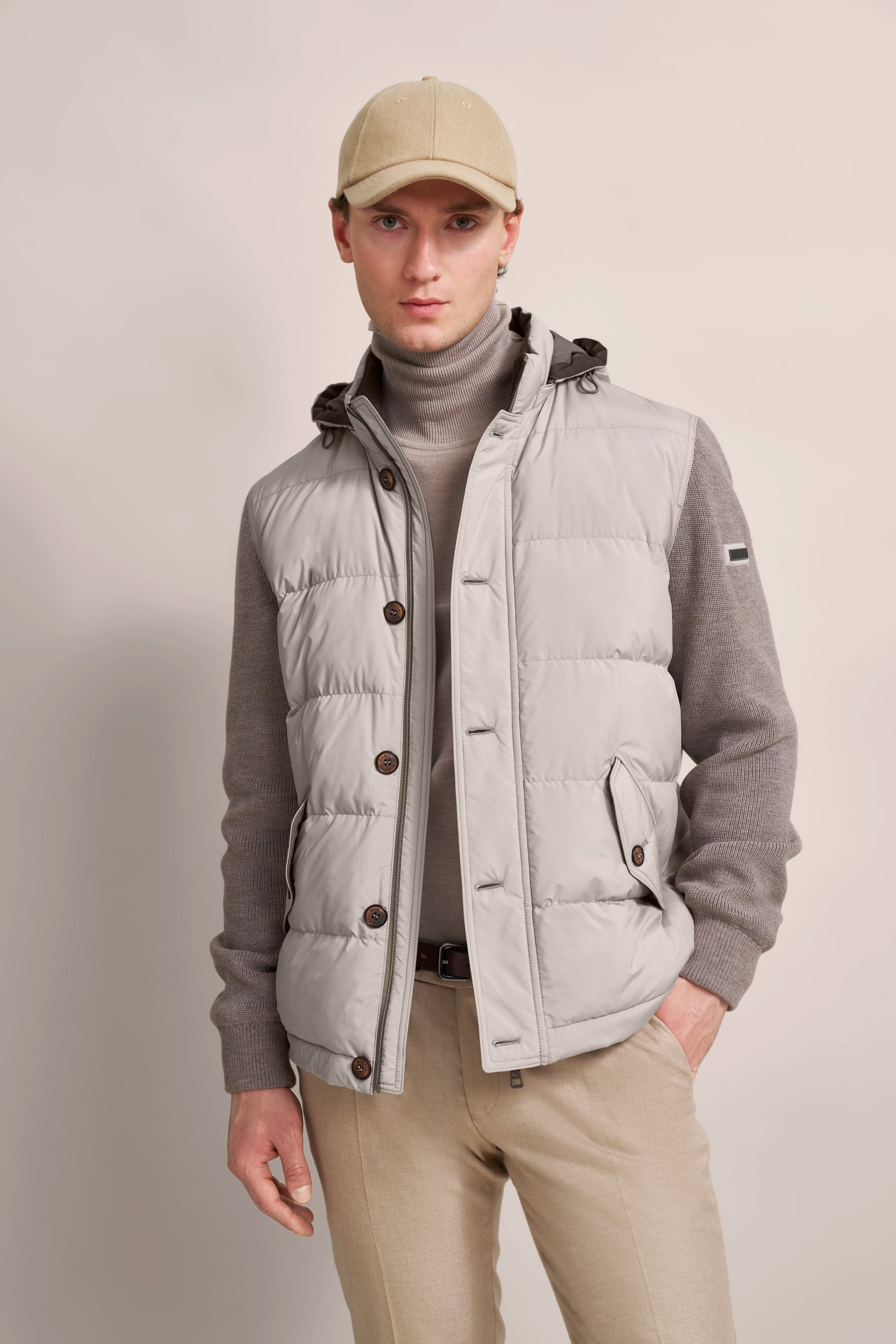 Model in beige vest, gray turtleneck sweater, beige pants, and cap. Casual, elegant pose.
